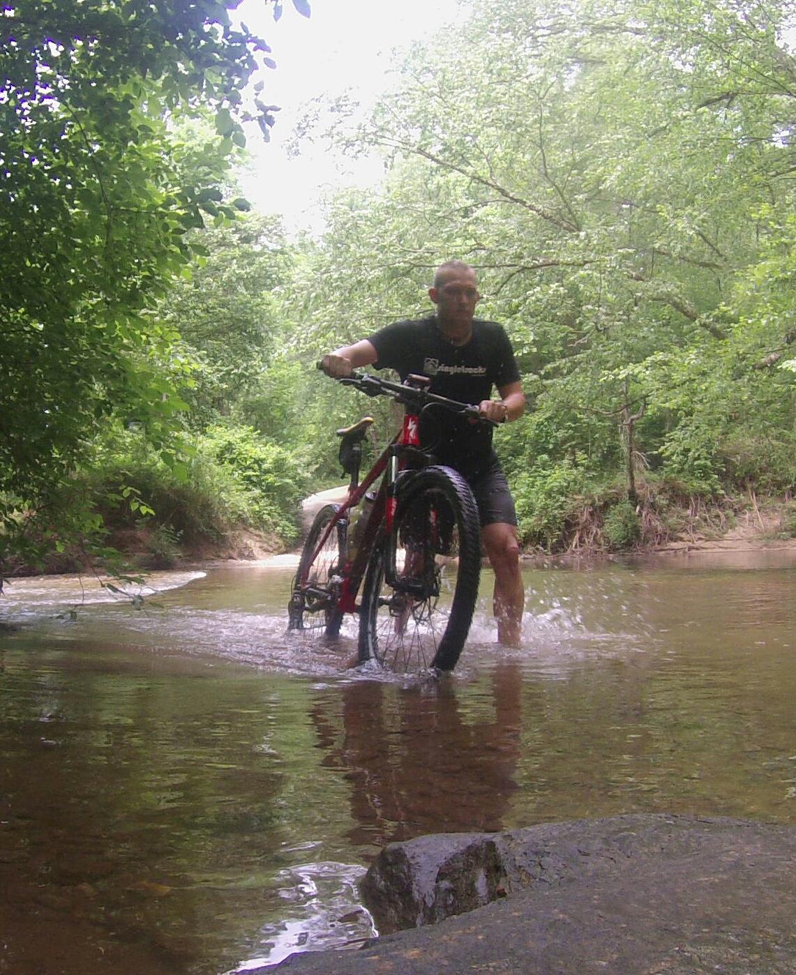 A person riding a mountain bike is wading through a shallow stream surrounded by lush green foliage. The cyclist is lifting the bike’s front wheel out of the water, creating ripples in the stream. The scene captures an adventurous outdoor experience in a natural setting. Dawson Forest mountain bike trail.