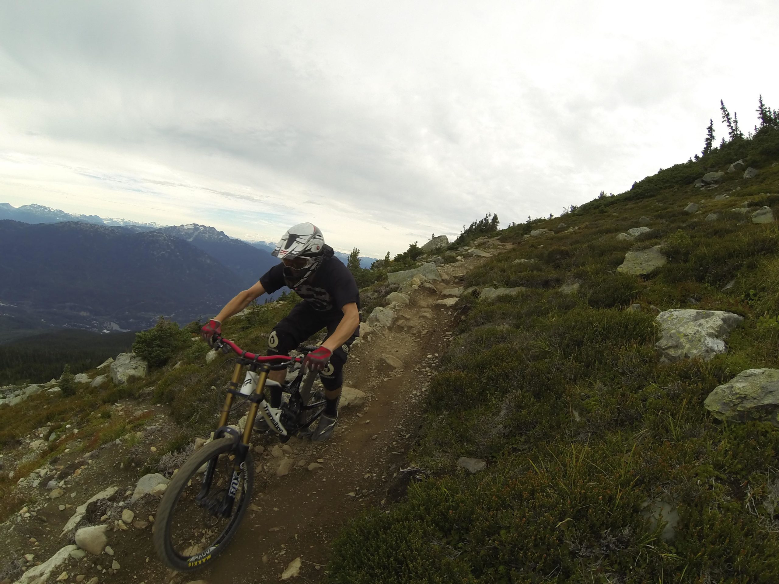 A mountain biker navigating a rocky trail, wearing a helmet and protective gear, with a scenic mountain range in the background under a cloudy sky. Top Of The World mountain bike trail.
