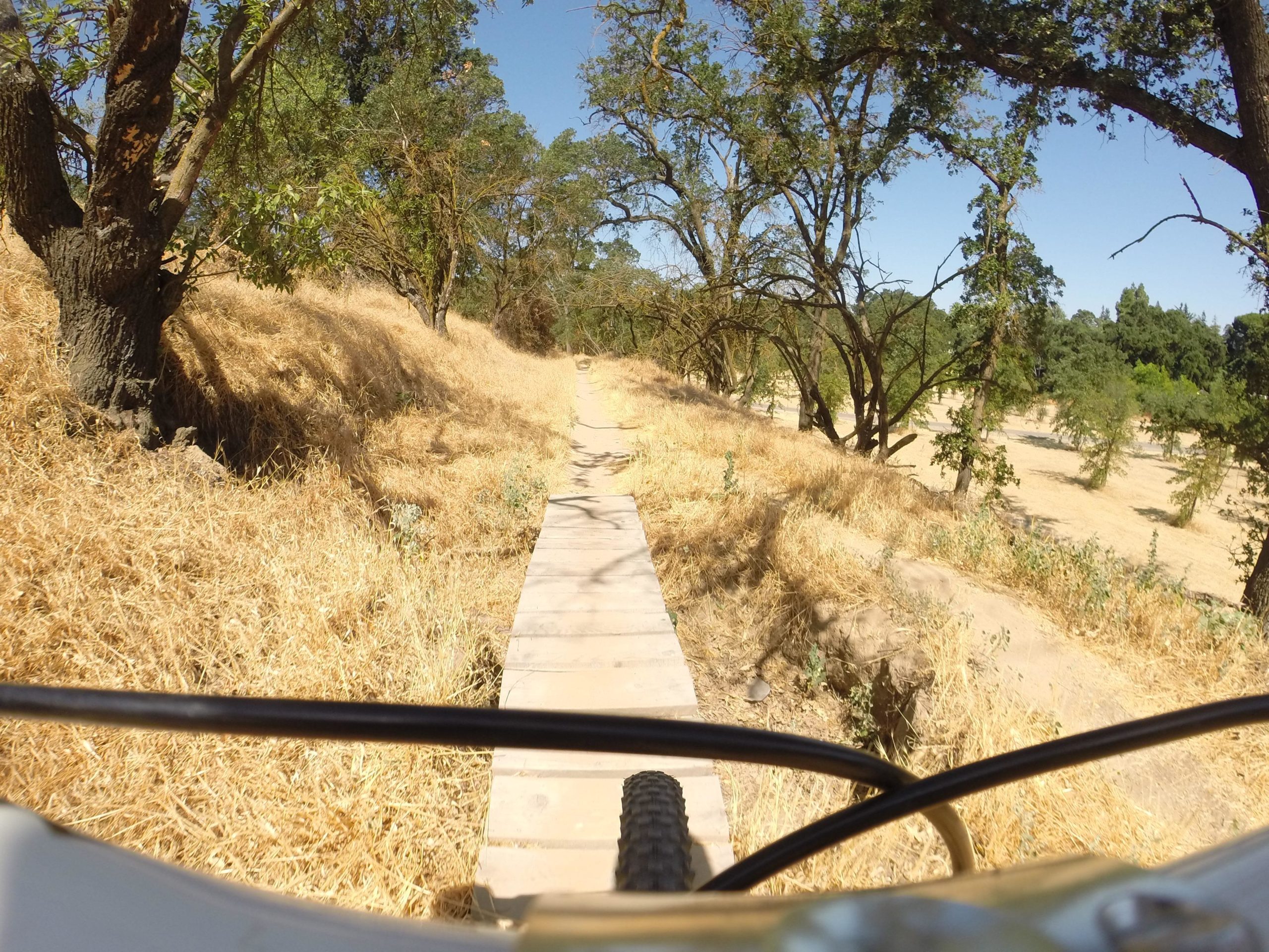 A wooden bike path winding through a dry, grassy area with trees on either side under a clear blue sky. The perspective is taken from the front of a bicycle, showcasing the trail ahead. Dry Creek Trail mountain bike trail.