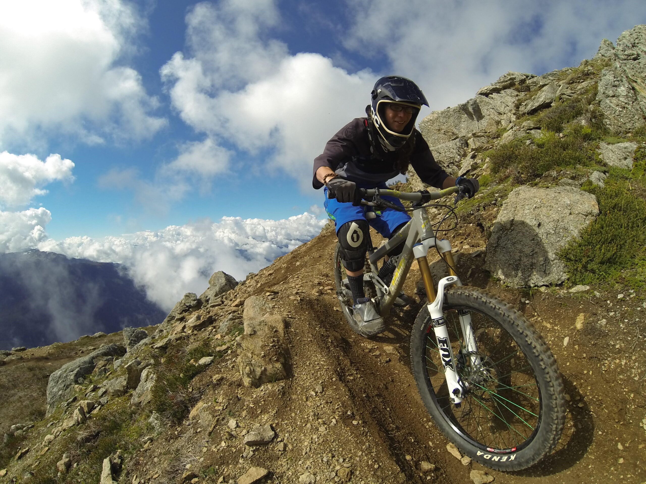 A mountain biker in protective gear descends a rocky trail on a mountainside, surrounded by clouds and a clear blue sky. The terrain is rugged, with visible rocks and dirt leading down the slope. The biker's focused expression and posture indicate an excitement for the ride. Top Of The World mountain bike trail.