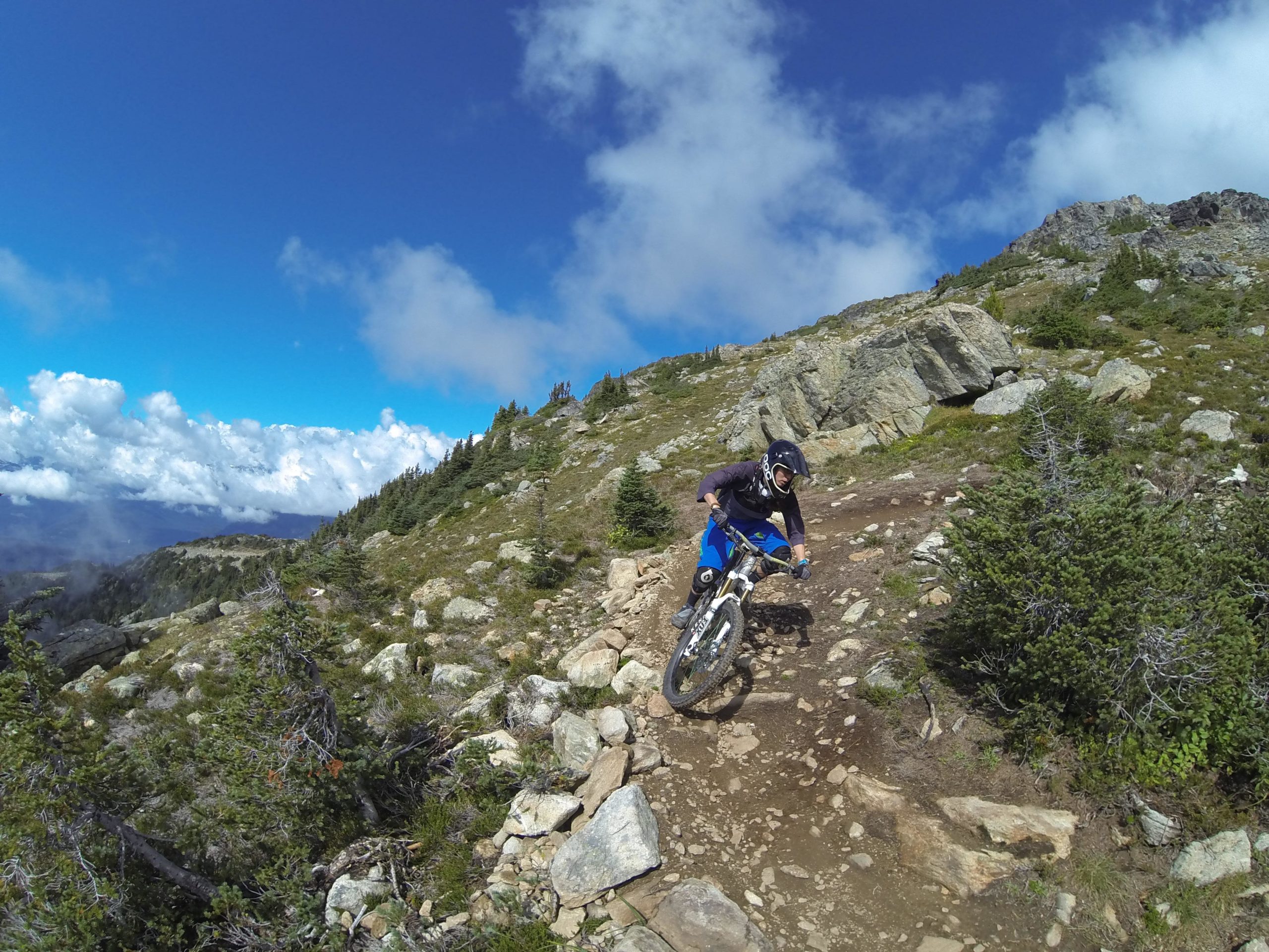 A mountain biker navigating a rocky trail on a sunny day, with clouds scattered in a blue sky. Lush greenery and trees frame the landscape, while rugged terrain is visible in the background. Top Of The World mountain bike trail.