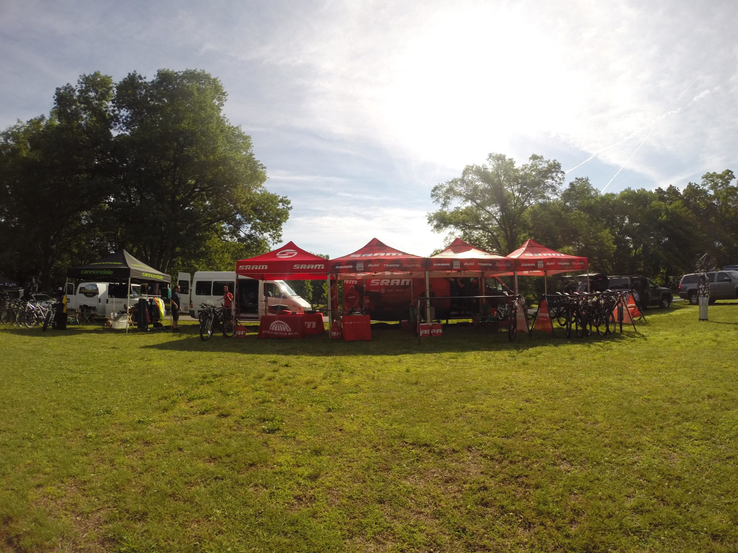 A vibrant outdoor scene featuring several tents set up for a biking event. The SRAM and Cannondale tents display their branding prominently, surrounded by bicycles and biking gear. In the background, a white van and a truck are visible, with green trees and a clear blue sky above, creating a lively atmosphere. Blue Mountain Reservation mountain bike trail.
