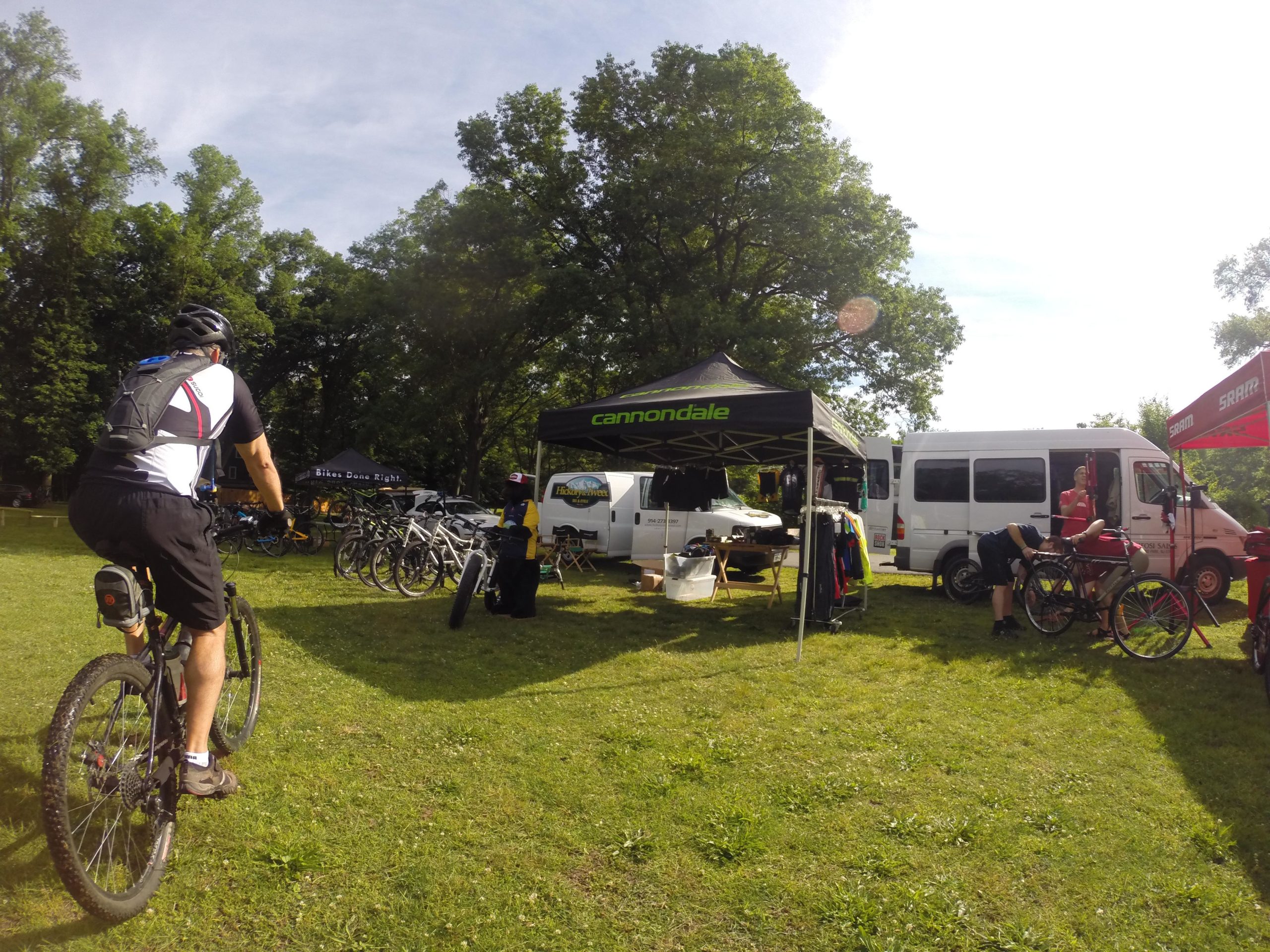 A cyclist in a black and white jersey rides towards a display area with tents and various bicycles. The scene is set in a grassy park surrounded by trees, with tents featuring cycling brands and vendors interacting with customers. Blue Mountain Reservation mountain bike trail.