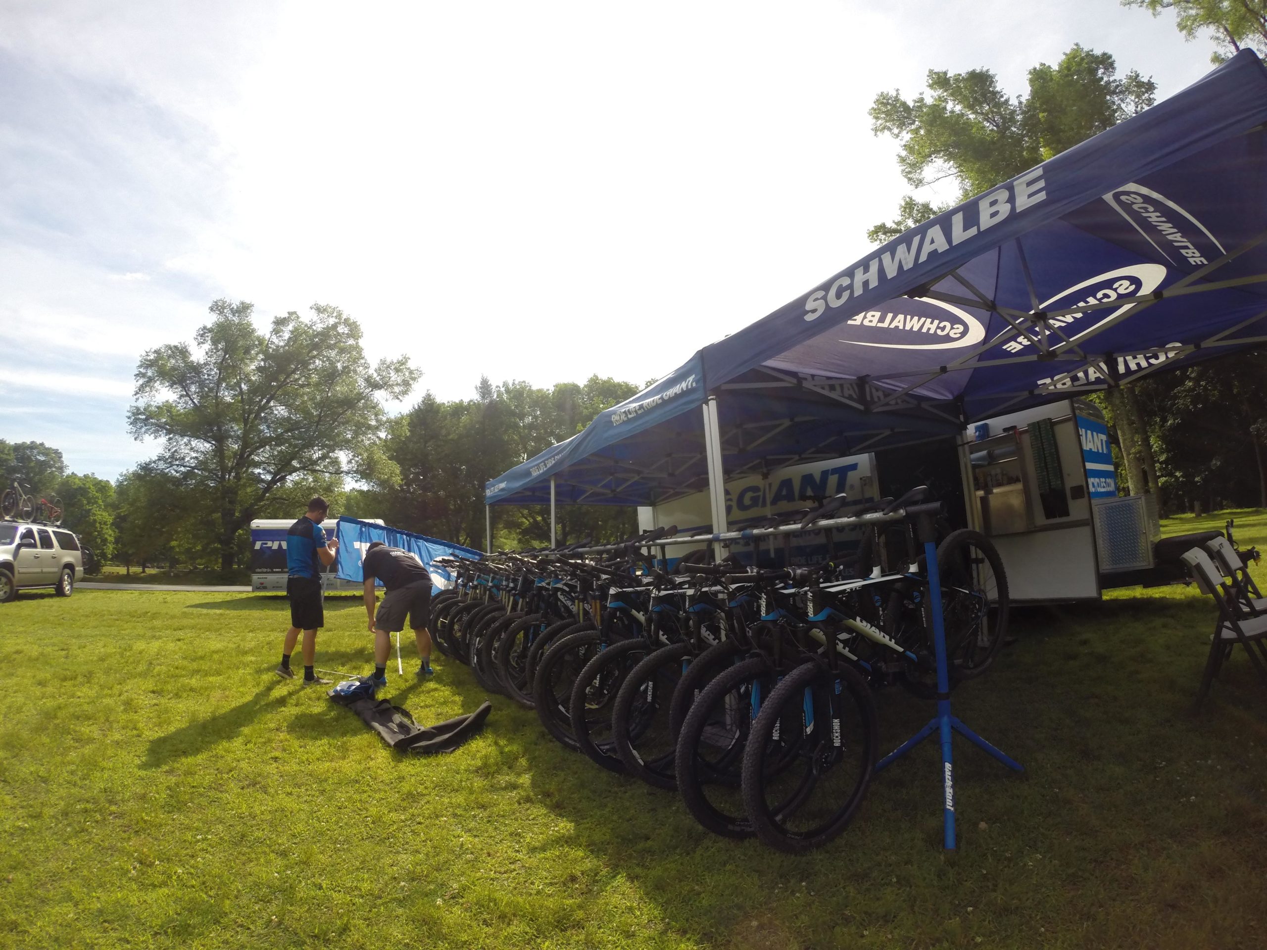 Two individuals set up equipment at a cycling event under blue tents displaying the brands Schwalbe and Giant. In the background, a row of mountain bikes is lined up on display, with a vehicle parked nearby on a green lawn surrounded by trees. The sky is clear and sunny. Blue Mountain Reservation mountain bike trail.