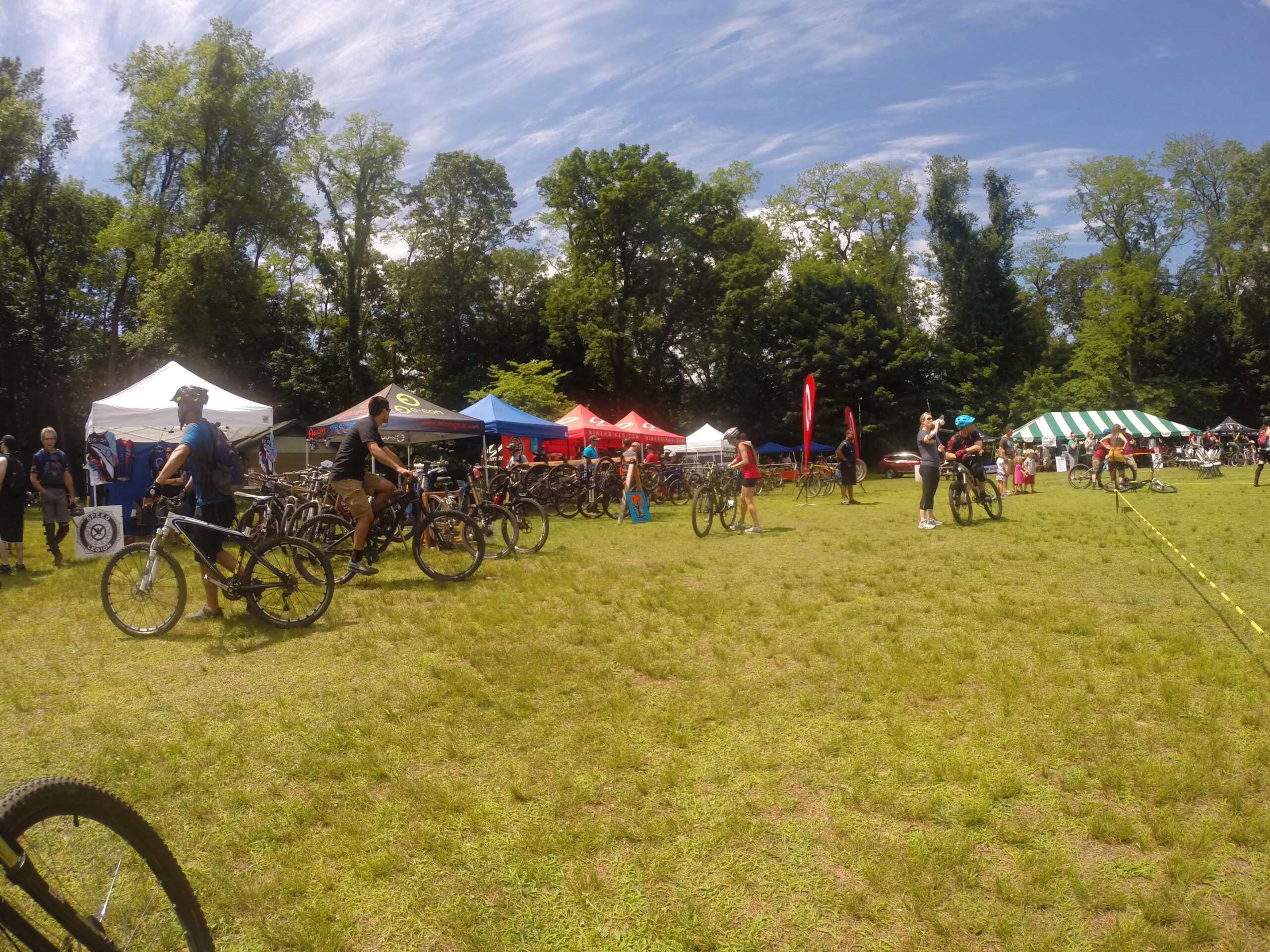 A lively outdoor scene at a mountain biking event, featuring participants in cycling gear among a crowd of bicycles. Tents and banners from various sponsors are set up in the background, with trees and a blue sky overhead. The grass is green and well-maintained, indicating an active, energetic atmosphere. Blue Mountain Reservation mountain bike trail.