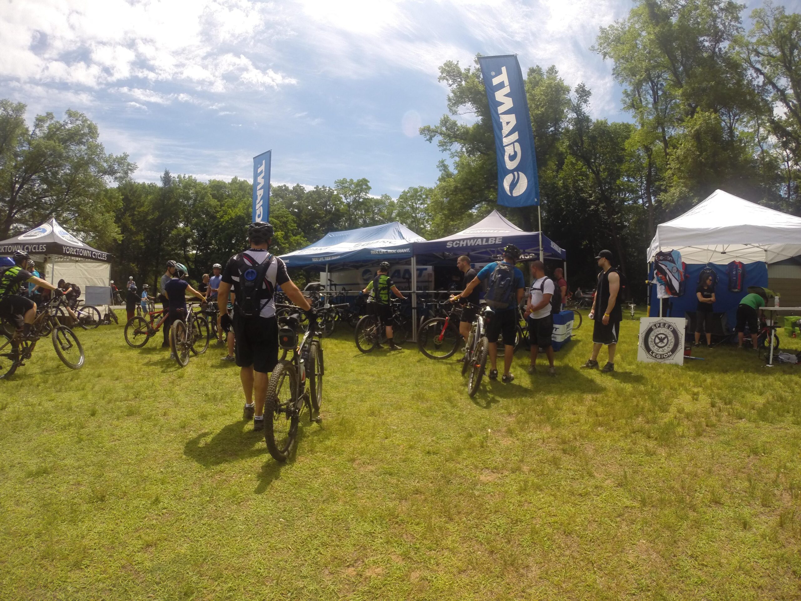 A group of cyclists gathered at a mountain biking event, with several tents displaying brand logos in the background. Participants are seen interacting, standing beside their bikes on a grassy field under a partly cloudy sky. The atmosphere is vibrant, showcasing the community and enthusiasm for cycling. Blue Mountain Reservation mountain bike trail.