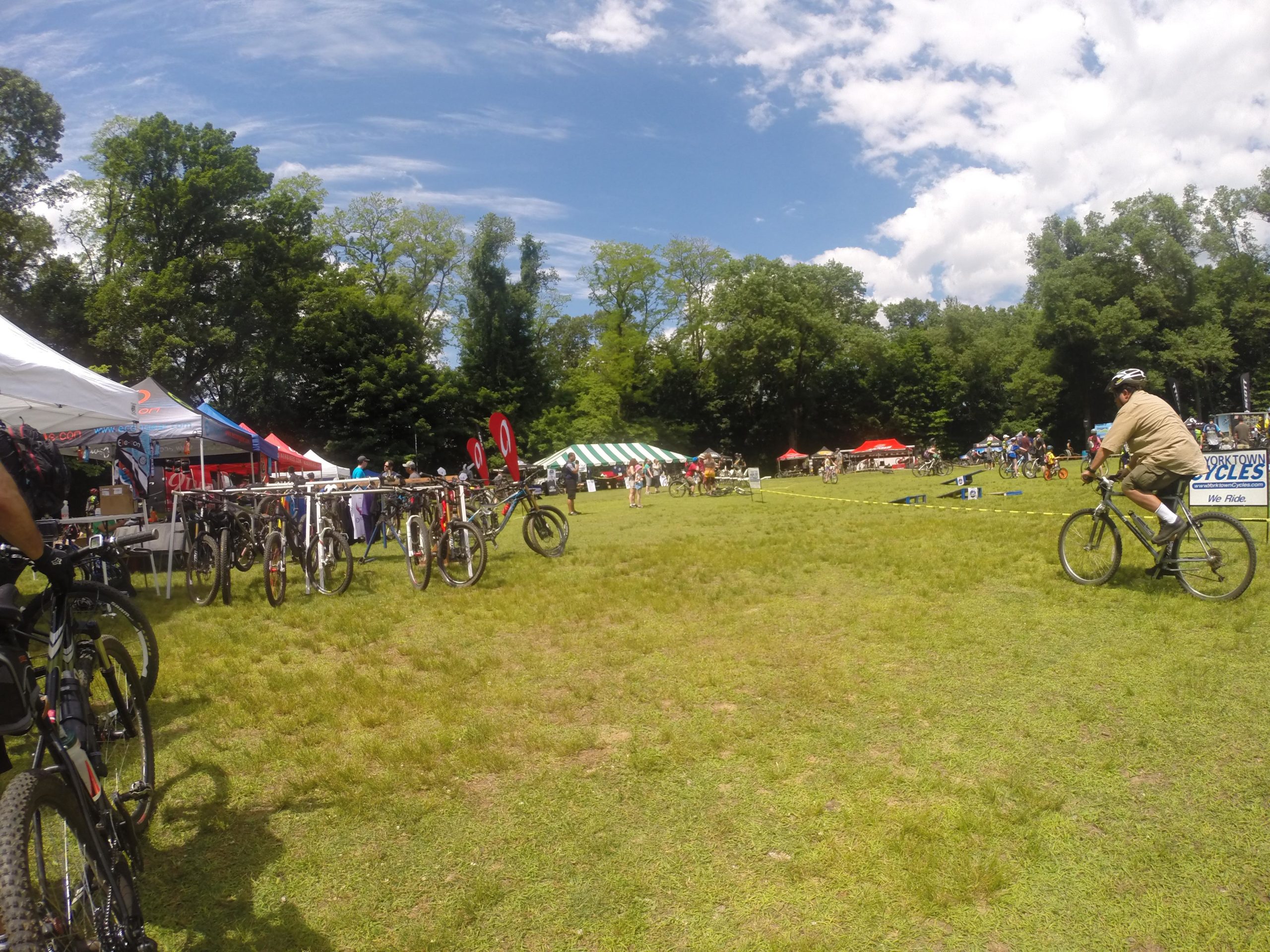 An outdoor scene at a cycling event featuring tents and booths, with various bicycles on display. Participants are engaged in activities on a grassy field under a clear blue sky with scattered clouds. A cyclist is seen riding in the foreground, while others explore the event in the background. Blue Mountain Reservation mountain bike trail.