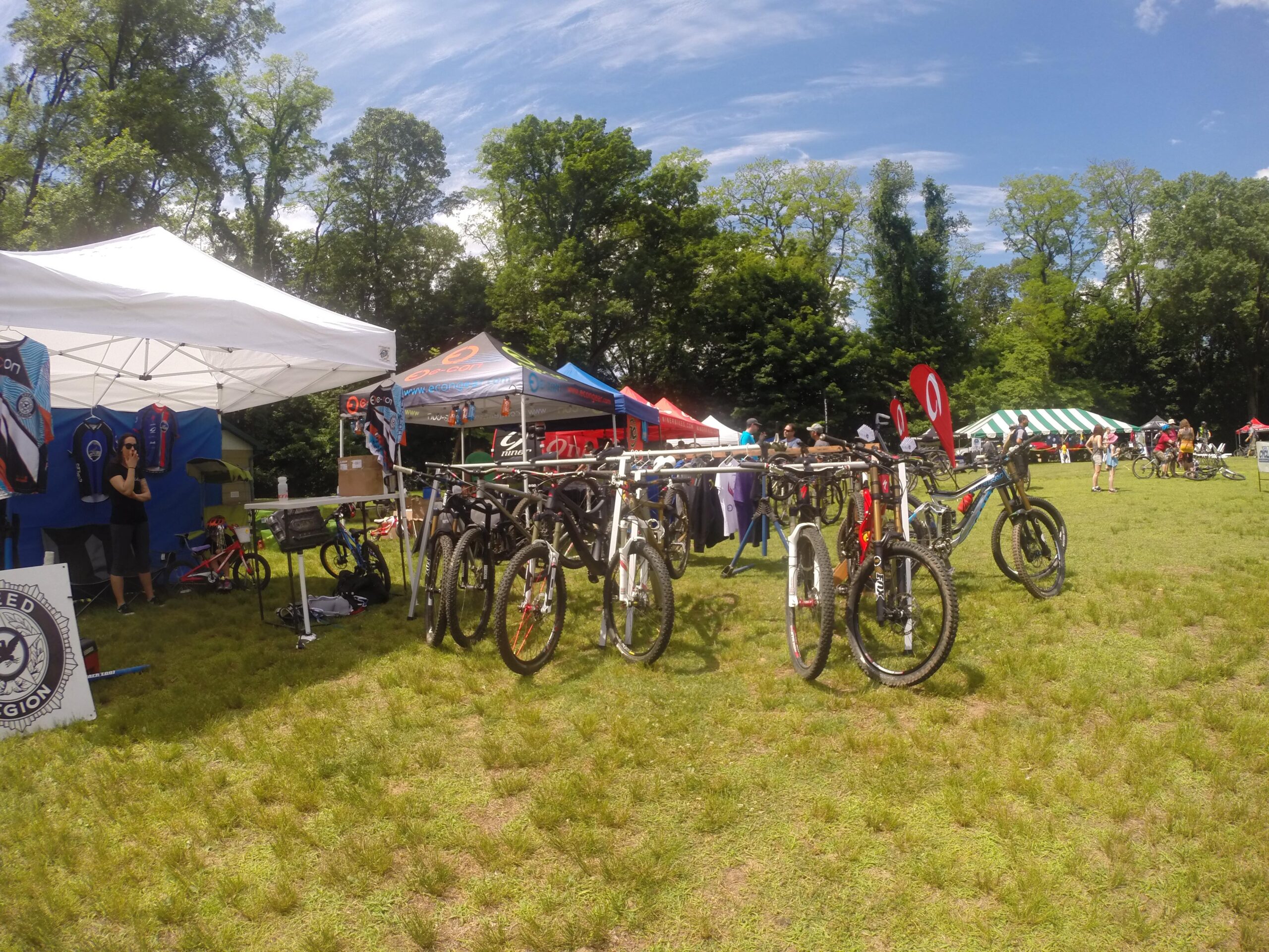 A vibrant outdoor event showcasing various mountain bikes under white tents. In the foreground, several mountain bikes are displayed on a rack, with a woman standing beside a booth featuring cycling apparel. Colorful tents in the background host additional vendors and spectators enjoying the day, surrounded by green grass and trees under a blue sky. Blue Mountain Reservation mountain bike trail.