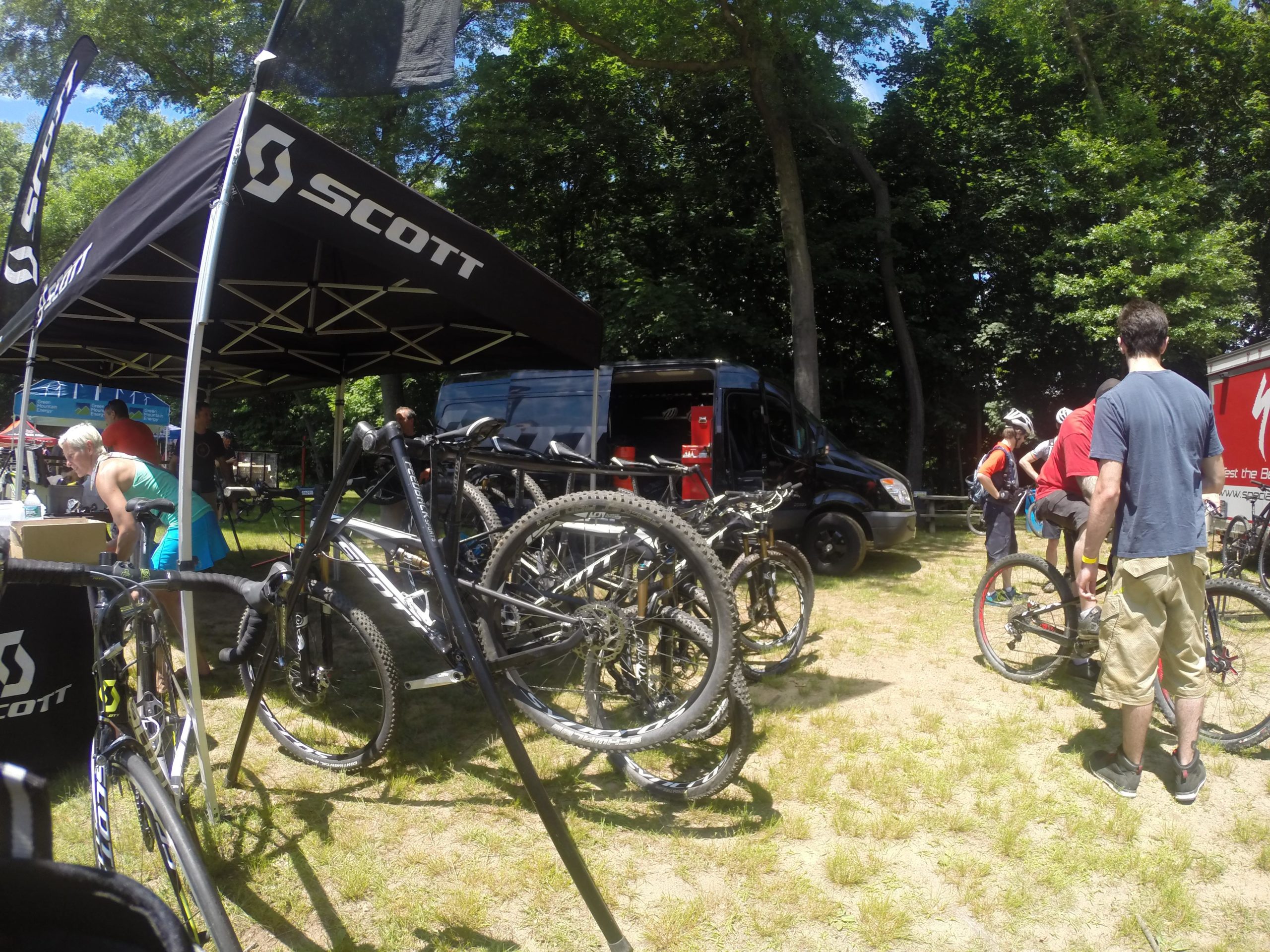 Image of a bicycle event with a canopy marked "Scott" set up in a wooded area. Several mountain bikes are lined up in the foreground, and individuals are engaged in activities related to biking, including examining bikes and preparing for rides. A black van is visible in the background, along with additional branding tents. The scene is vibrant and sunny, showcasing a lively outdoor atmosphere. Blue Mountain Reservation mountain bike trail.