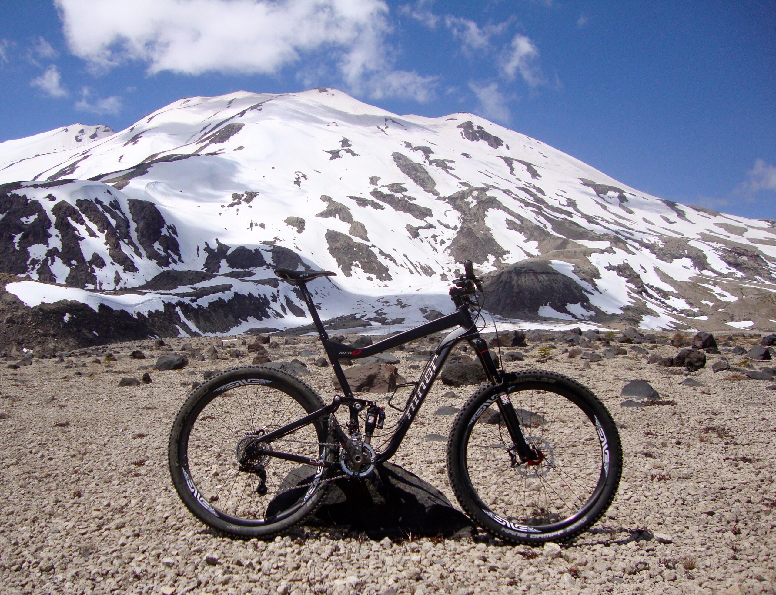 Niner W.F.O. 9: A black mountain bike is positioned on rocky terrain with a snow-capped mountain in the background under a partly cloudy sky.