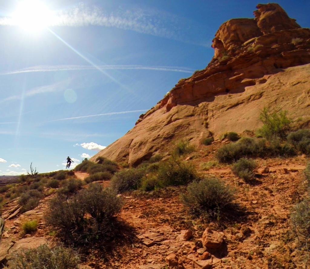 A cyclist rides along a rocky trail in a desert landscape, with sun shining brightly in a clear blue sky and dramatic rock formations in the background. Sparse vegetation and shrubs cover the foreground, highlighting the rugged terrain. Hymasa mountain bike trail.