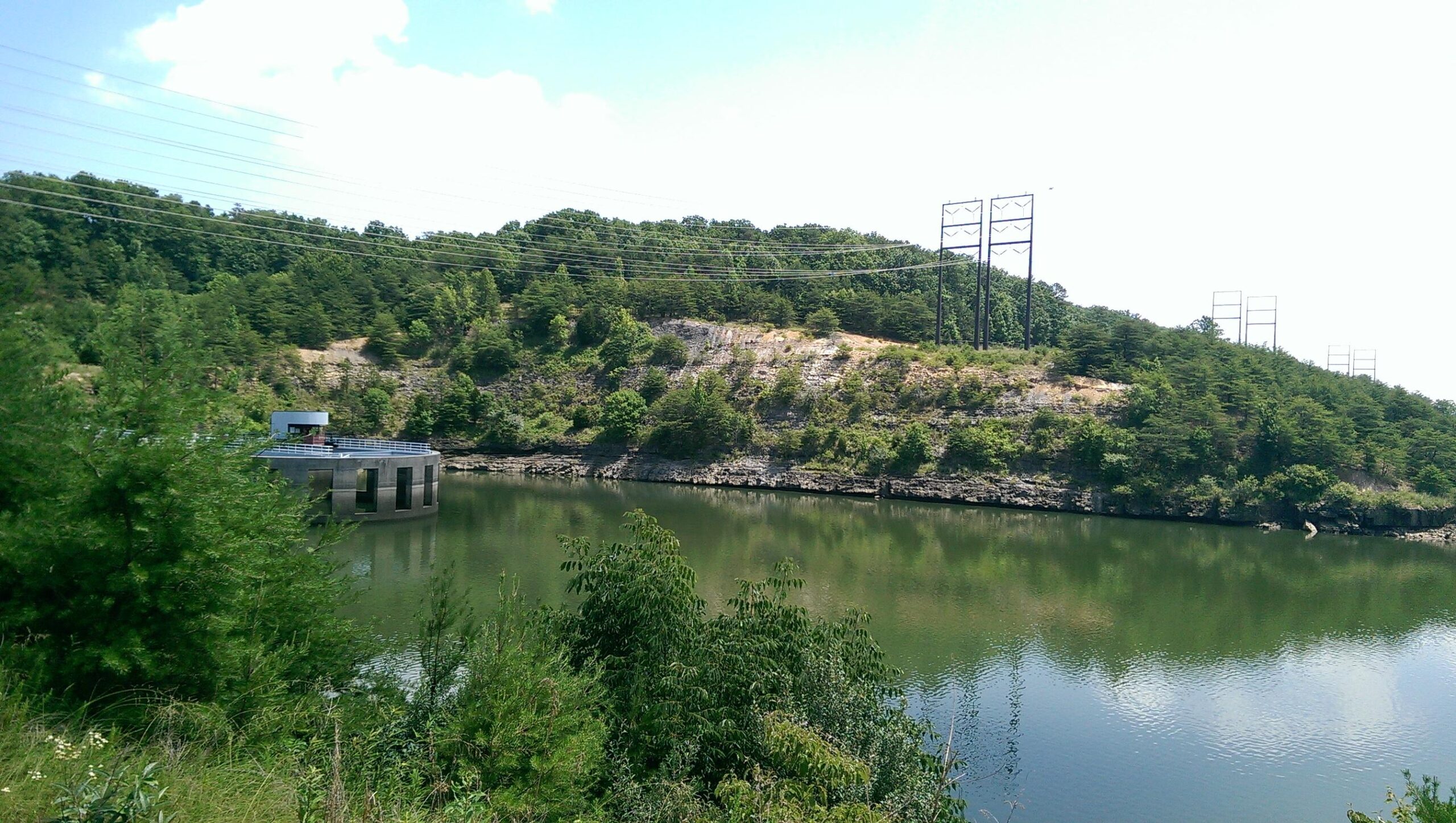 A serene landscape featuring a calm body of water surrounded by lush greenery and a wooded hillside. In the foreground, there are various shrubs and small trees. A concrete structure with a platform is visible on the water, and power lines stretch across the sky in the background. The scene is bathed in daylight, enhancing the vivid green of the trees and the reflections on the water's surface. Raccoon Mountain Trail Network mountain bike trail.