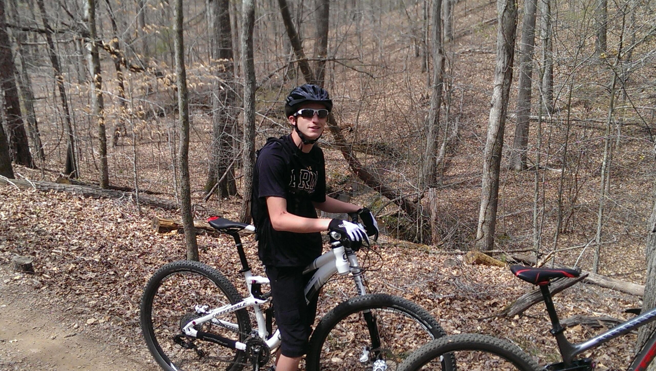 A young man wearing a black helmet and sunglasses stands beside a mountain bike on a dirt trail in a wooded area. The surroundings are composed of bare trees and fallen leaves, indicating early spring. The man is dressed in a black t-shirt and shorts, and he holds the bike's handlebars while looking at the camera. Another bike is in the background. Dwelling Loop mountain bike trail.