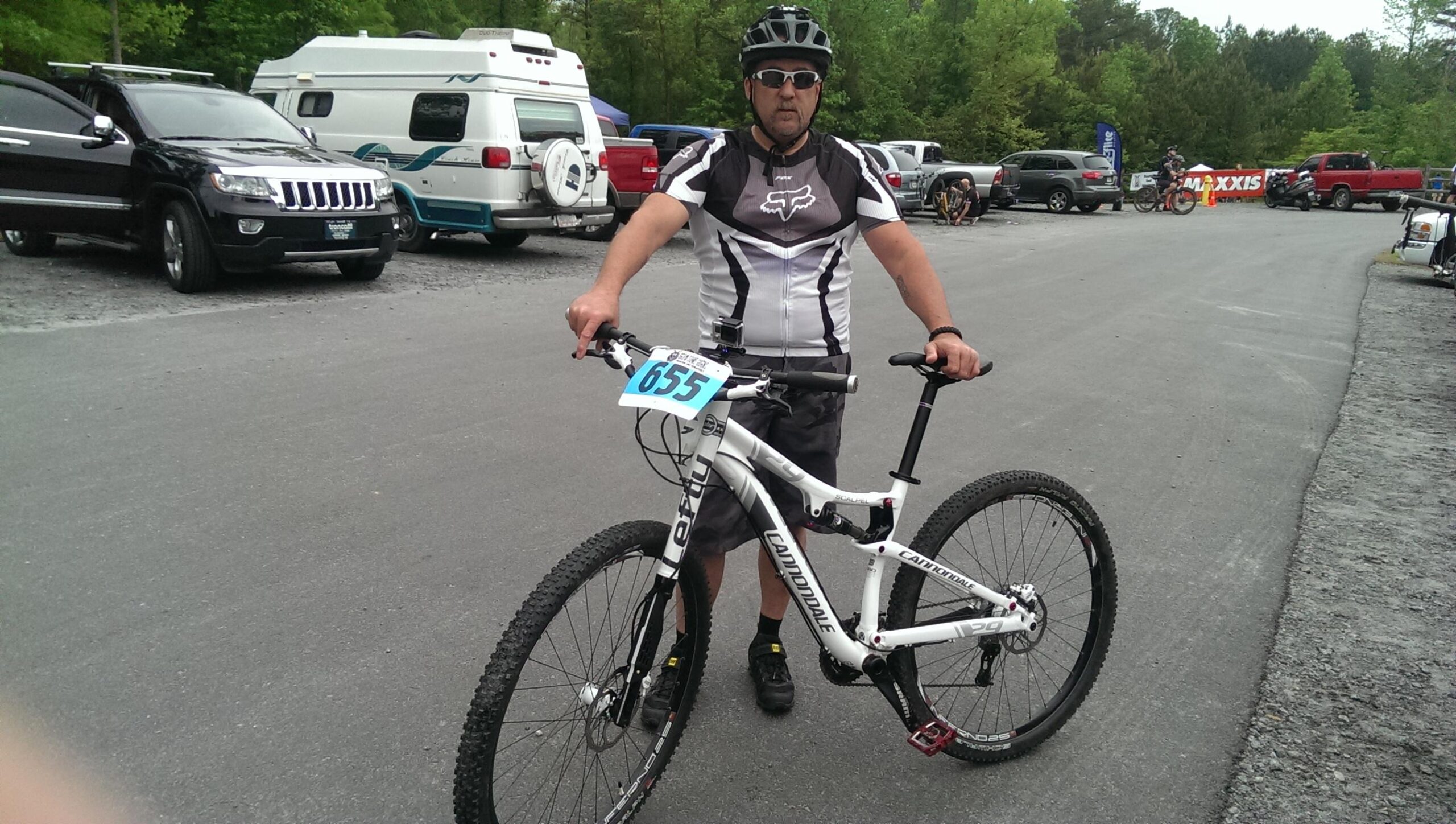 Cannondale Scalpel 29er 3: A man wearing a black and white cycling jersey and sunglasses stands next to a white mountain bike. The bike displays a race number (655) on it. In the background, various vehicles, including a black SUV and a camper van, are parked along a gravel lot surrounded by greenery. The scene appears to be set at a biking event or competition.