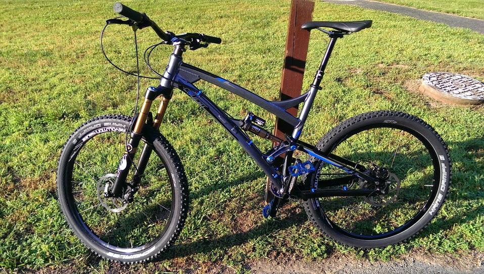 Transition Covert 27.5: A blue and black mountain bike with front suspension, positioned on green grass. The bike features wide tires, a sleek frame, and a raised seat post, with a wooden post in the background and a circular manhole cover partially visible.