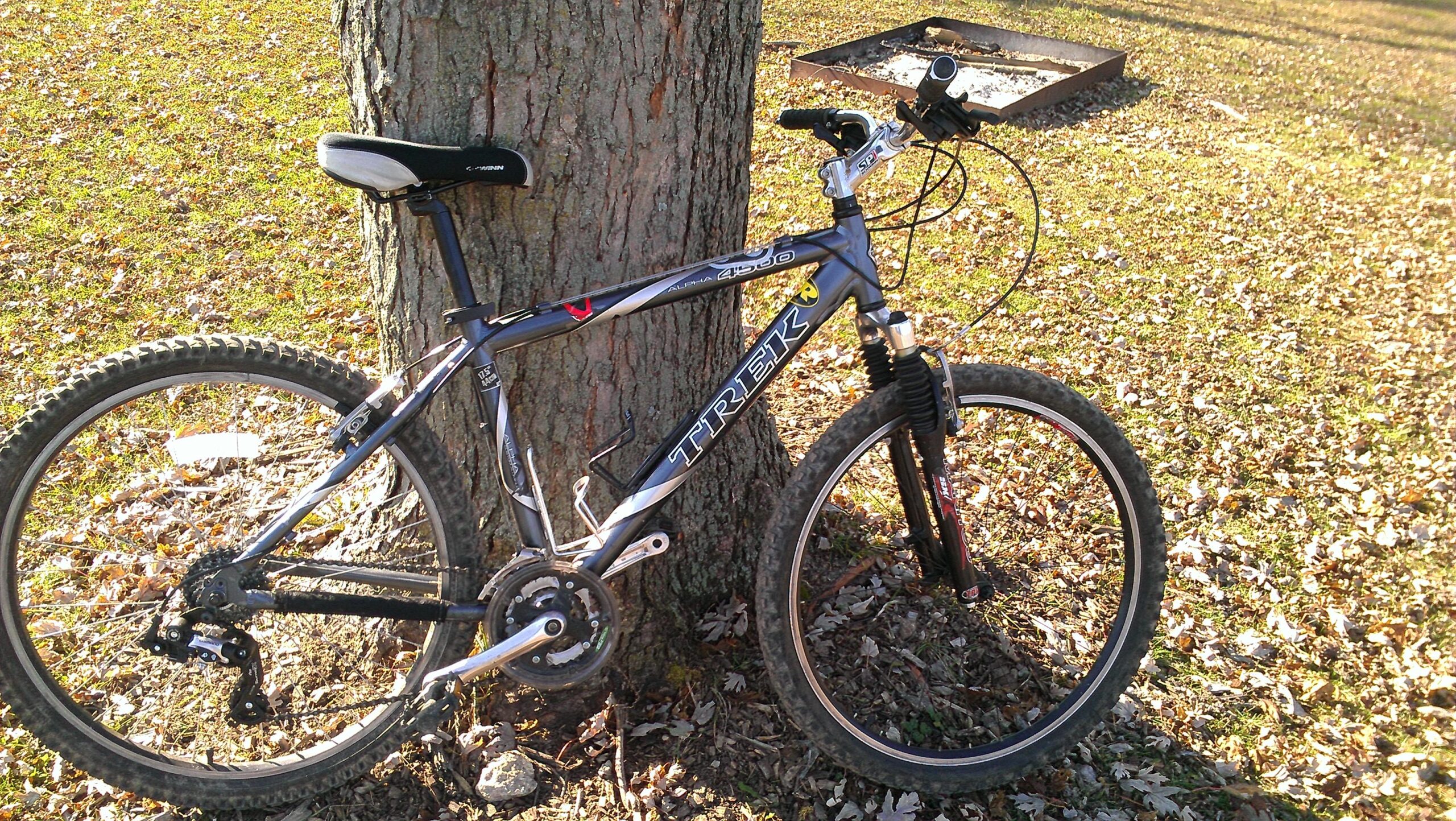 Trek 4500: A mountain bike leaning against a tree, with fallen leaves scattered on the ground in a grassy area. The bike has a silver and black frame with visible gears and tires designed for off-road riding.