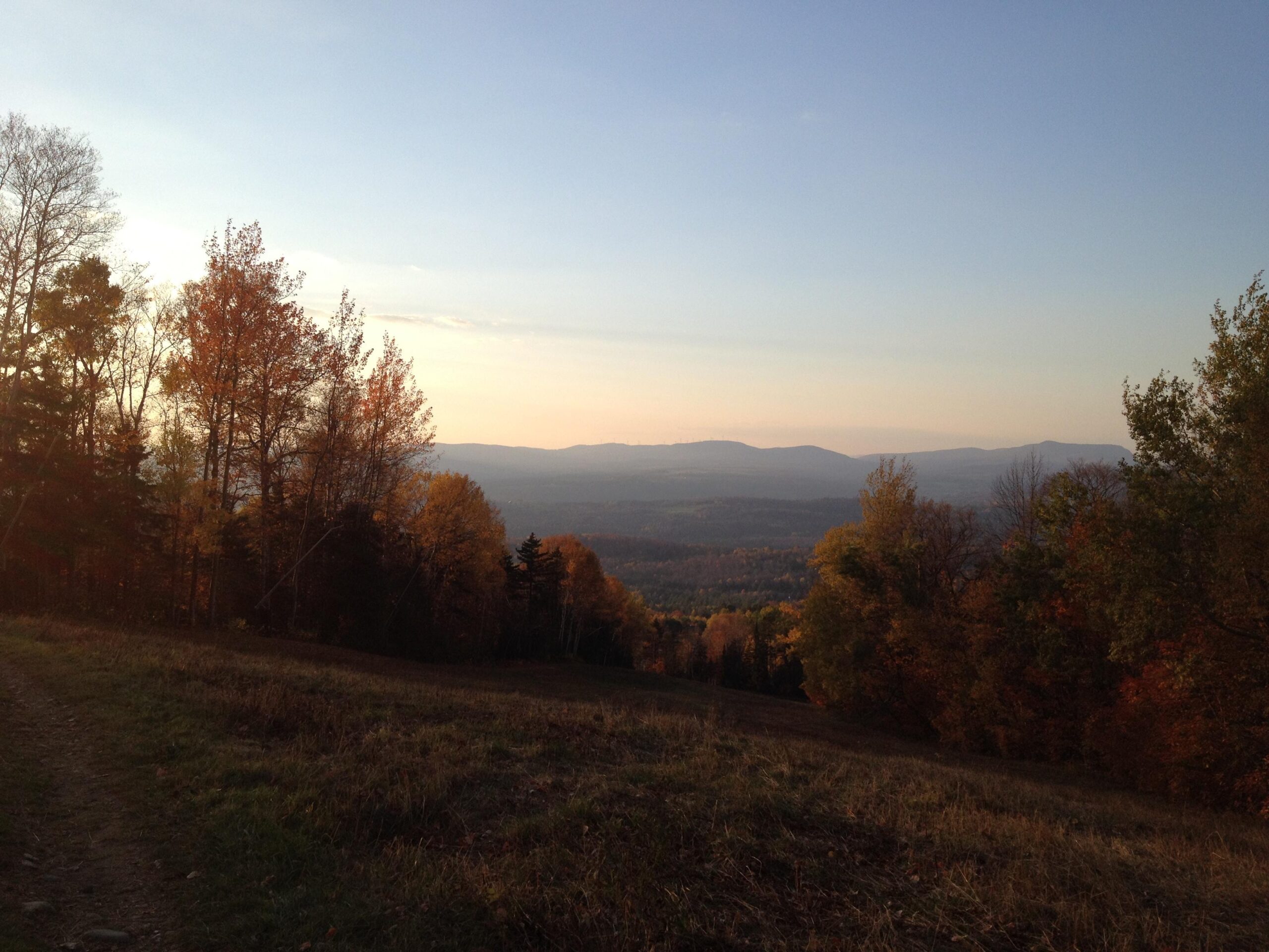 A scenic view of a mountainous landscape during autumn, featuring trees with orange and yellow foliage. The foreground has a grassy area leading to a dirt path, while the background showcases rolling hills and a clear sky as the sun sets. Kingdom Trails mountain bike trail.