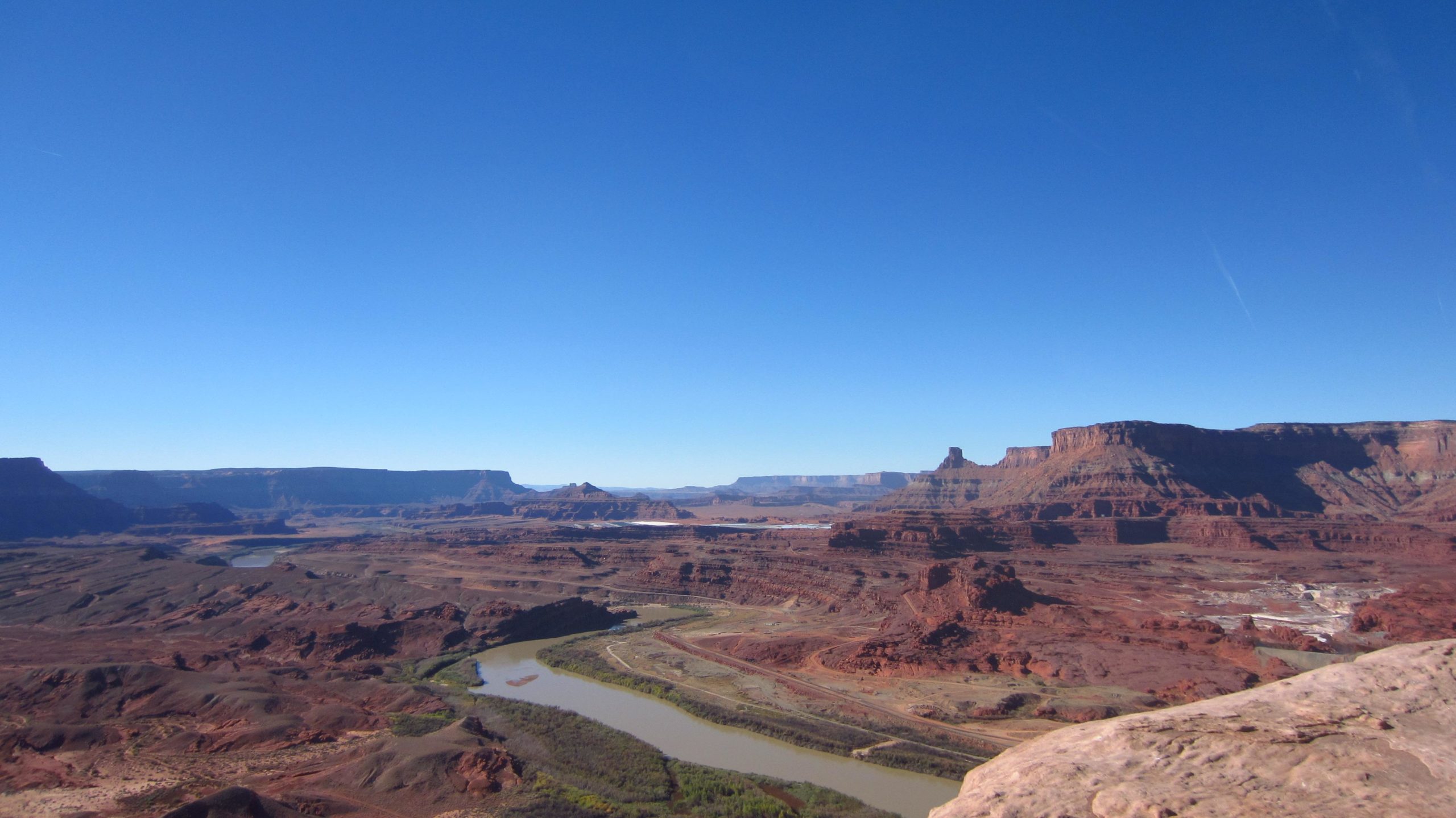 A panoramic view of rugged red rock formations and canyons under a clear blue sky, with a winding river flowing through the landscape. Amasa Back Trail mountain bike trail.