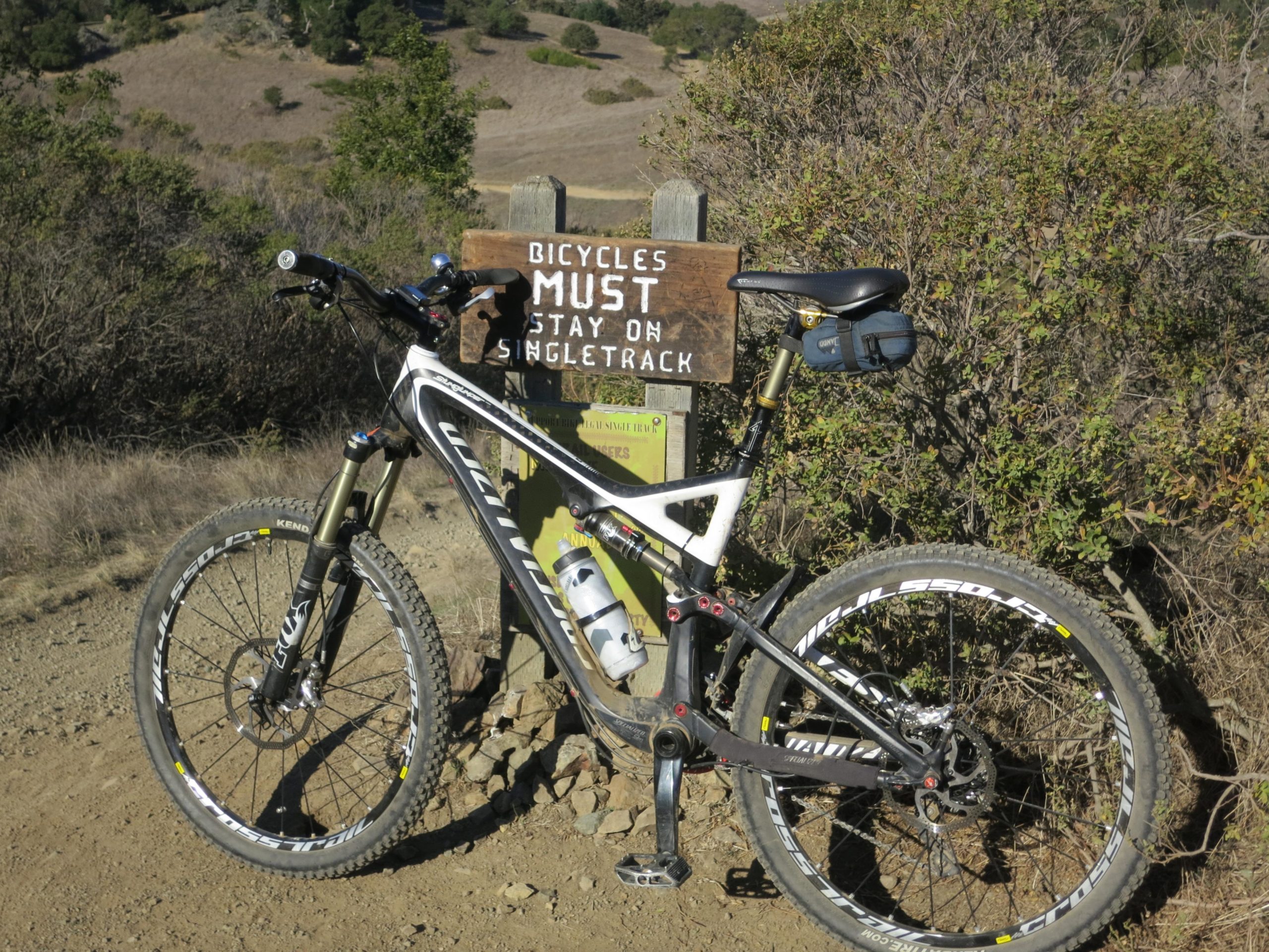 A mountain bike parked next to a wooden sign that reads "Bicycles Must Stay on Singletrack." The background features a hilly landscape with sparse vegetation and dry grass. The sun is shining, indicating a clear day. Camp Tamarancho mountain bike trail.
