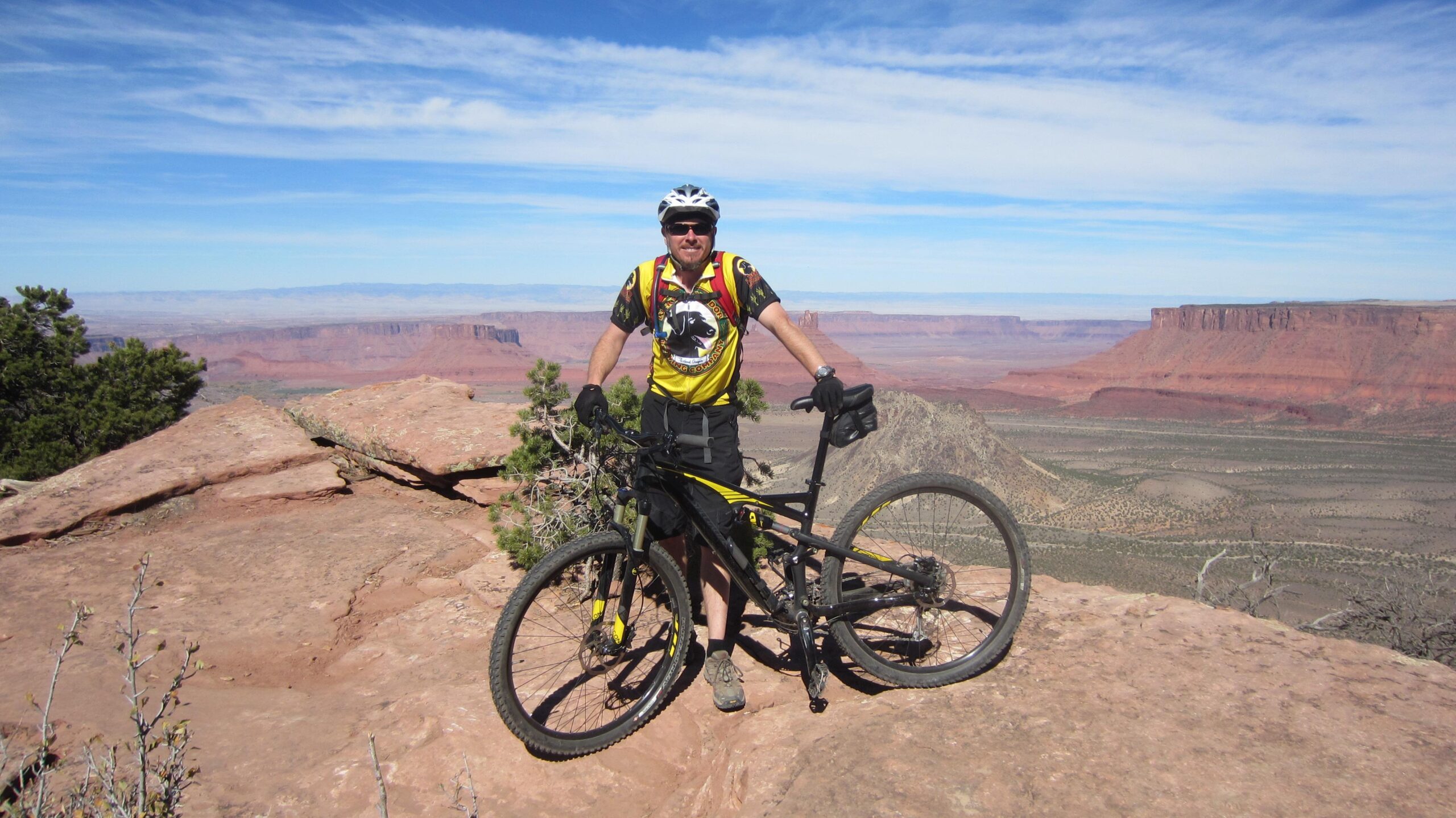 A person wearing a yellow and black cycling jersey and helmet stands next to a mountain bike on a rocky outcrop, with a vast landscape of red rock formations and blue skies in the background. The Whole Enchilada mountain bike trail.