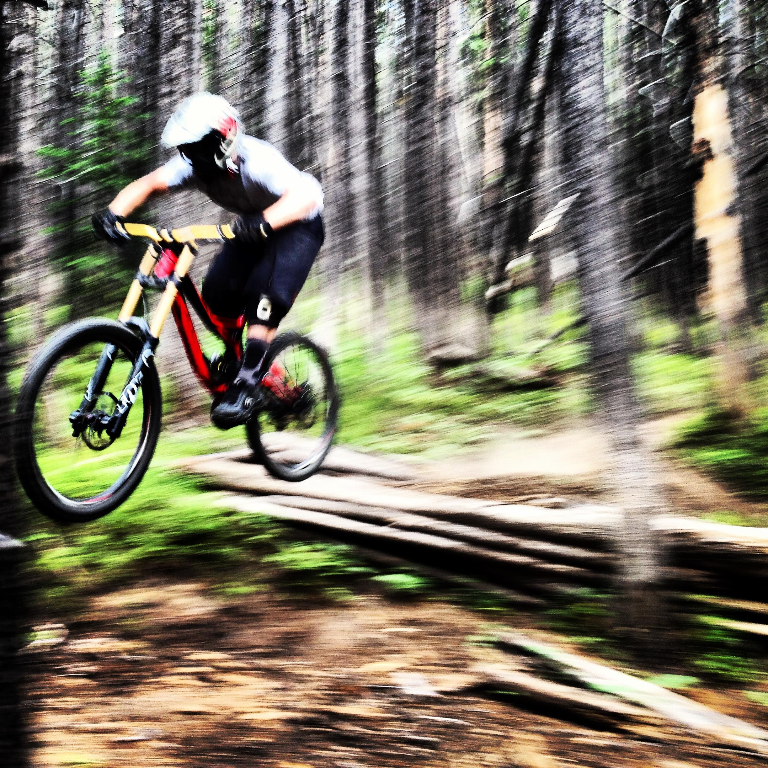 A mountain biker mid-jump on a trail, set against a blurred backdrop of trees, suggesting high speed and action. The biker is wearing a helmet and protective gear, with a focus on the bike's colorful frame. Vail Mountain Bike Park mountain bike trail.