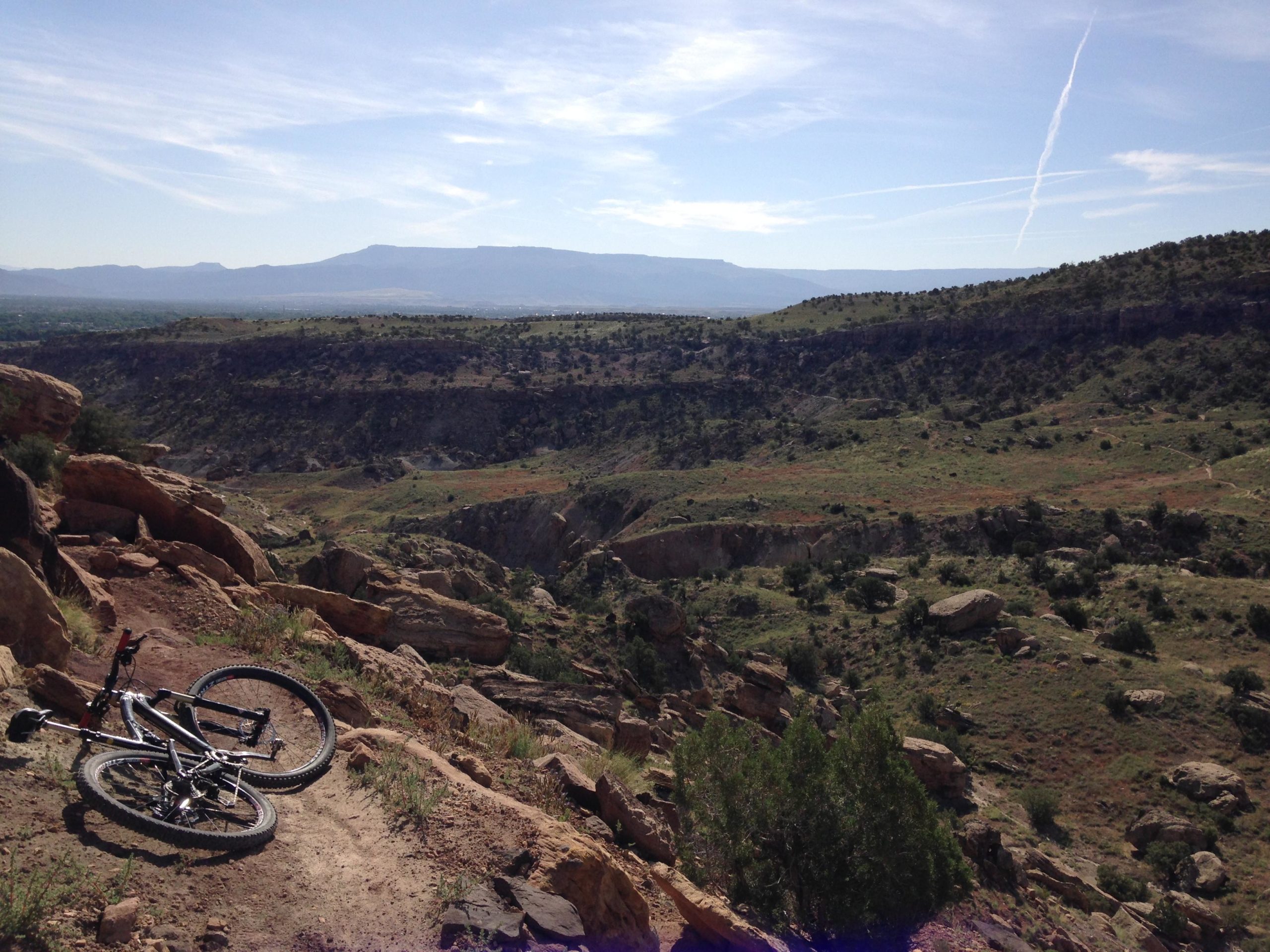 A mountain bike rests on a rocky trail overlooking a vast, expansive landscape of rolling hills and canyons under a blue sky with wispy clouds. Gunny Loop mountain bike trail.