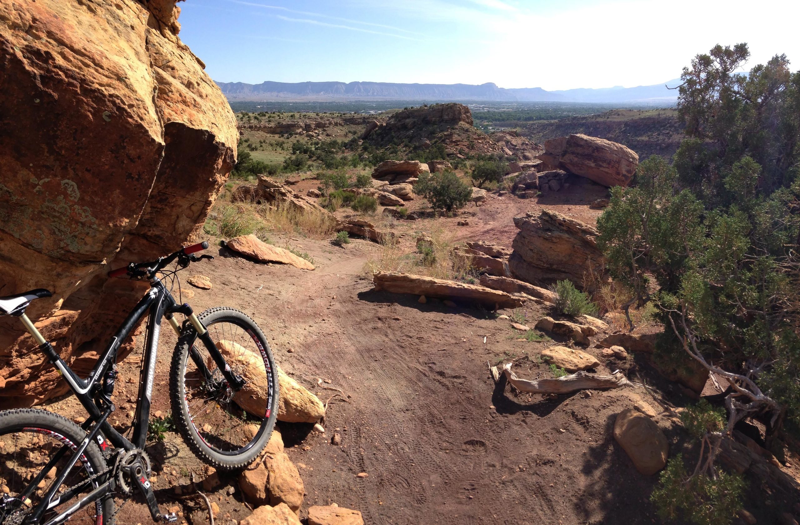 A mountain bike parked beside large boulders on a dirt trail, with a scenic view of rolling hills and distant mountains under a clear blue sky. Sunlight illuminates the rocky landscape, highlighting patches of greenery amidst the earthy tones of the terrain. Gunny Loop mountain bike trail.