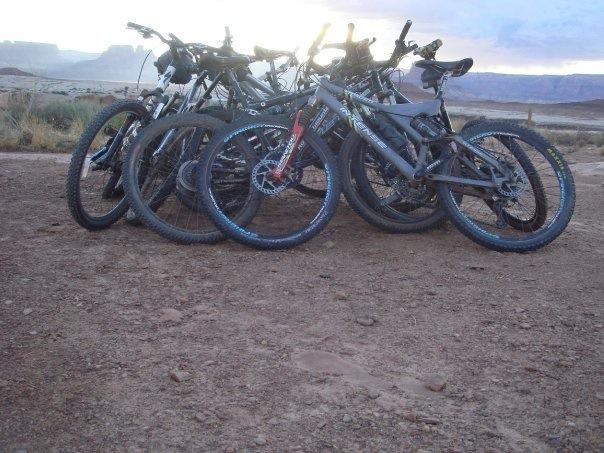 A group of mountain bikes is parked in a rugged outdoor setting, with a dirt surface and distant rocky cliffs in the background. The bikes, primarily in black and gray colors, are arranged in a pile with visible tires and handlebars, indicating a recreational outing in nature. The sky features soft clouds illuminated by sunlight. White Rim Trail mountain bike trail.