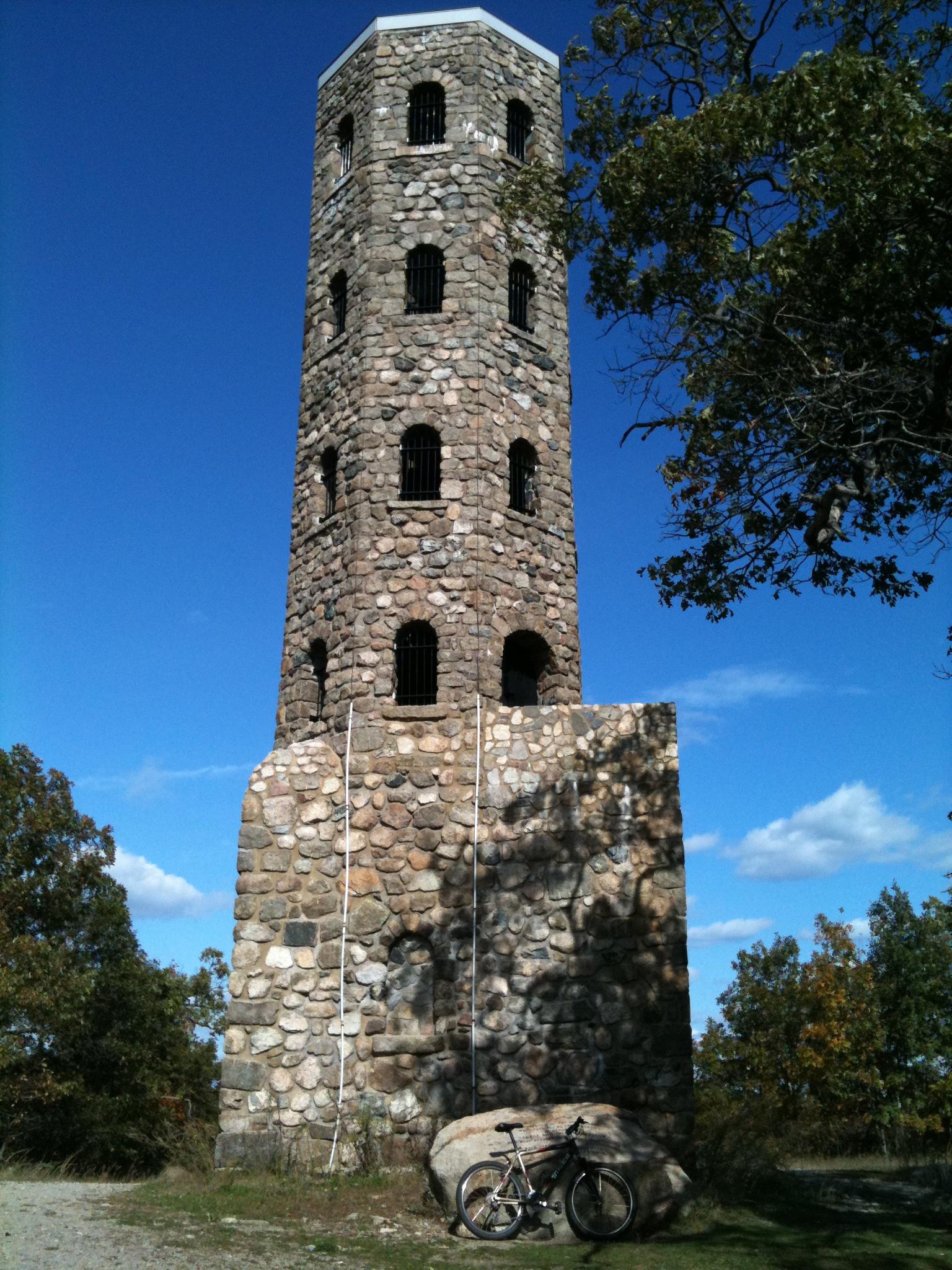 A tall stone tower with arched windows stands against a clear blue sky, surrounded by trees. A bicycle leans against a large rock in the foreground, suggesting a scenic outdoor location. The tower's rugged texture and construction materials are visible in the sunlight. Lynn Woods Reservation mountain bike trail.
