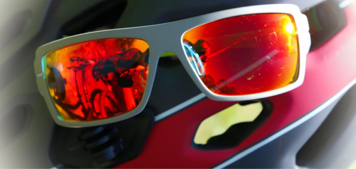 A close-up image of sunglasses with orange tinted lenses resting on a black and red bike helmet, with reflections of bicycles visible in the lenses. Apex Park mountain bike trail.