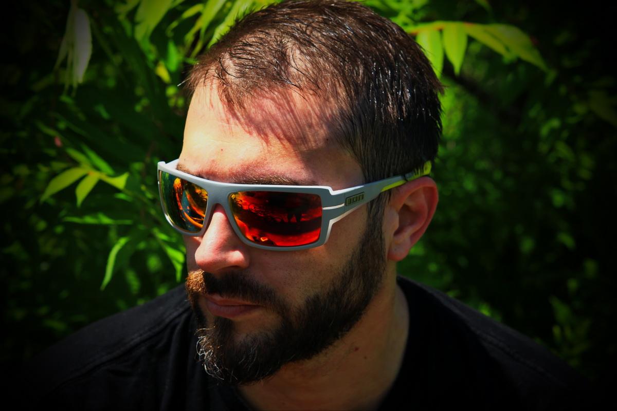 A man with a beard is wearing stylish sunglasses with red lenses, sitting outdoors in front of green foliage. The lighting highlights his facial features and the reflective surface of his sunglasses. He appears contemplative, with a serious expression. Don Valley mountain bike trail.