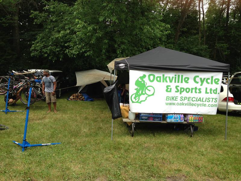 A tent labeled "Oakville Cycle & Sports Ltd." featuring a bicycle graphic, set up in a grassy area surrounded by trees. A person stands nearby, while bicycles are visible on racks and a car is parked in the background. The scene suggests a cycling event or pop-up shop.