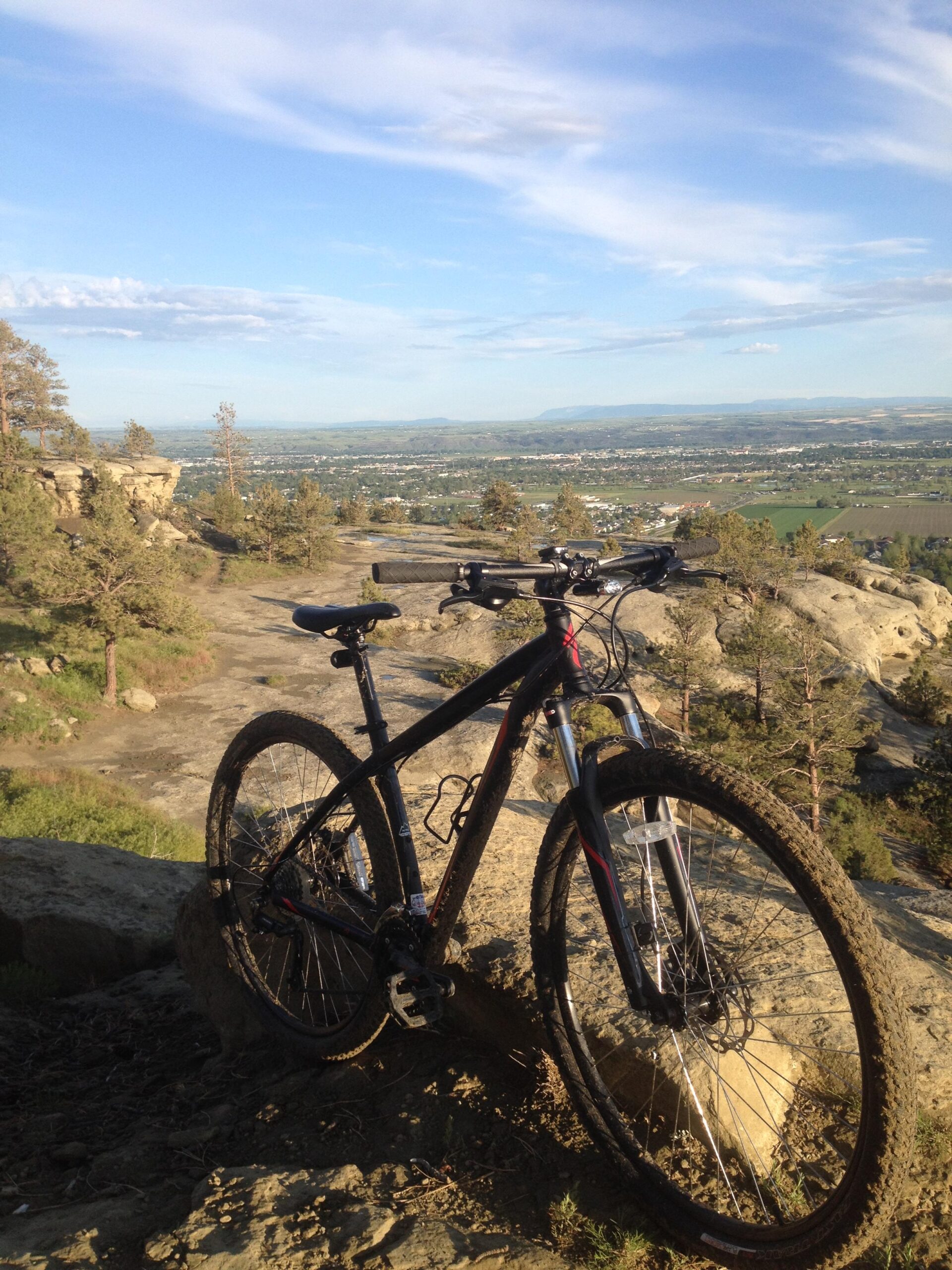 Specialized Hardrock Disc 29: A mountain bike resting on rocky terrain, overlooking a scenic valley filled with greenery and distant mountains under a clear blue sky with wispy clouds.