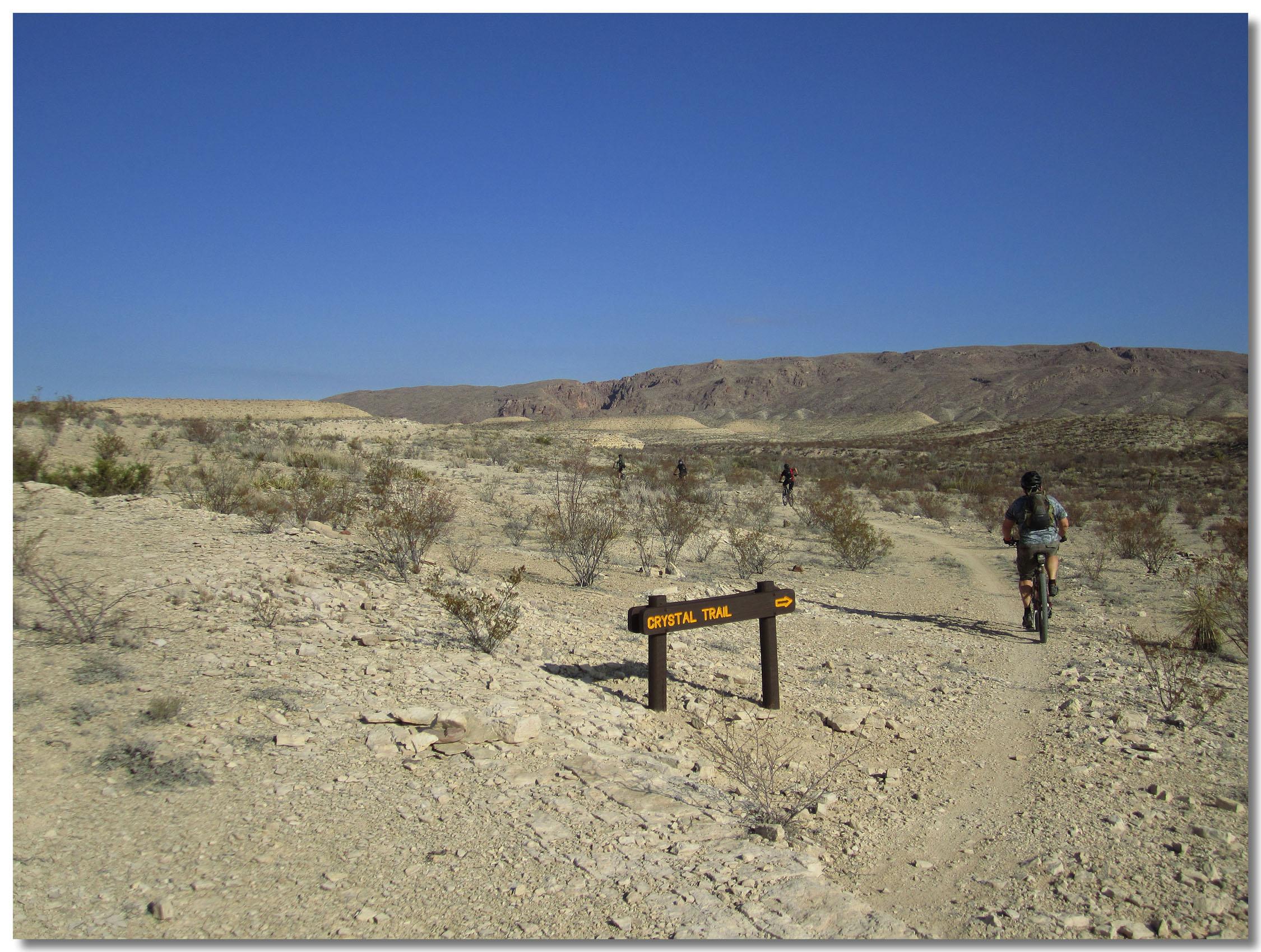 A dirt trail labeled "Crystal Trail" with directional signage pointing right, set in a desert landscape. In the background, rolling hills and a clear blue sky are visible. Two mountain bikers can be seen riding along the trail, surrounded by sparse vegetation and rocky terrain. Big Bend State Park mountain bike trail.