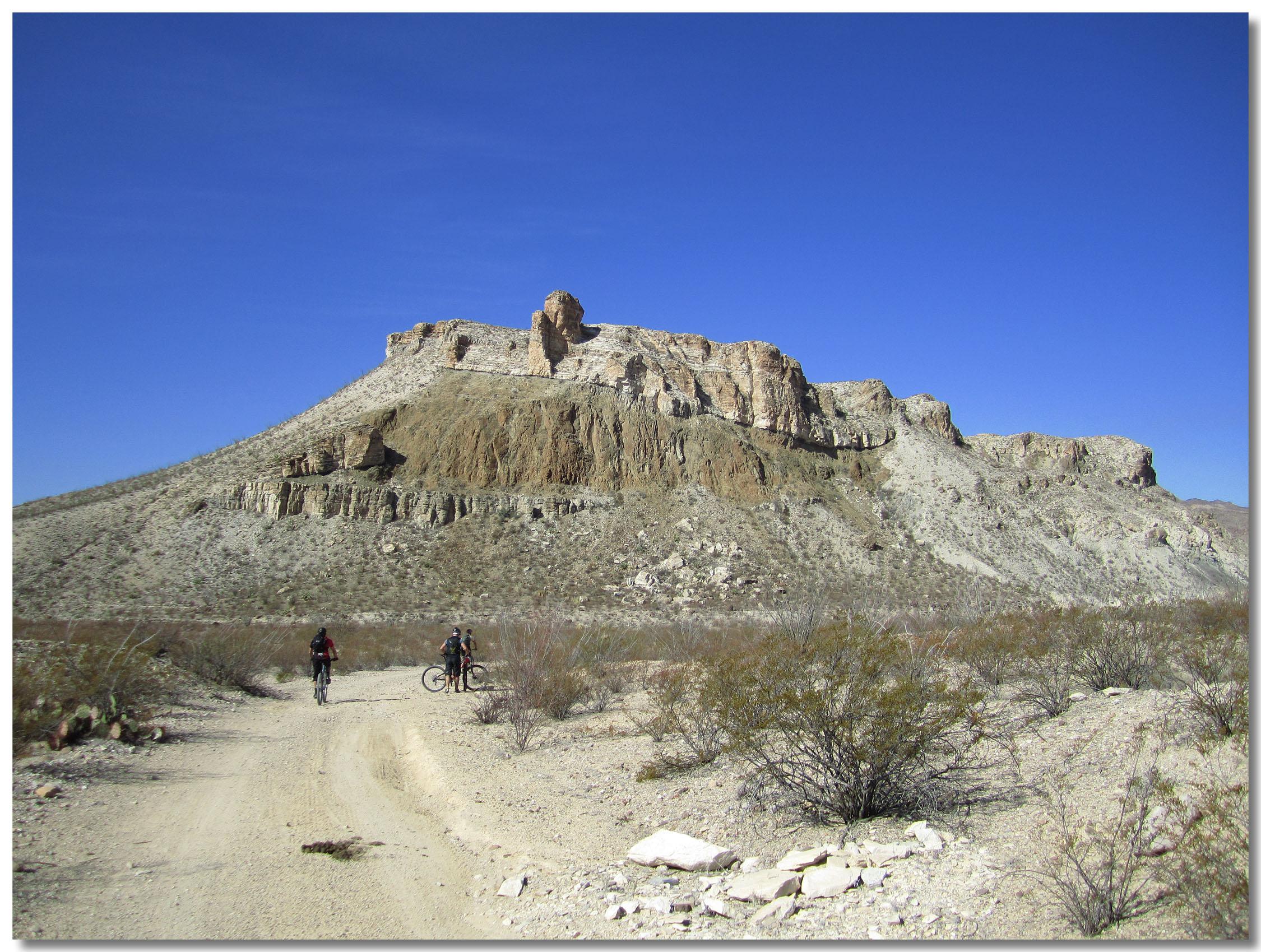 Two mountain bikers on a dirt path in a desert landscape, with a prominent rocky hill in the background under a clear blue sky. Sparse vegetation and rocky terrain surround the cyclists. Big Bend Ranch State Park mountain bike trail.