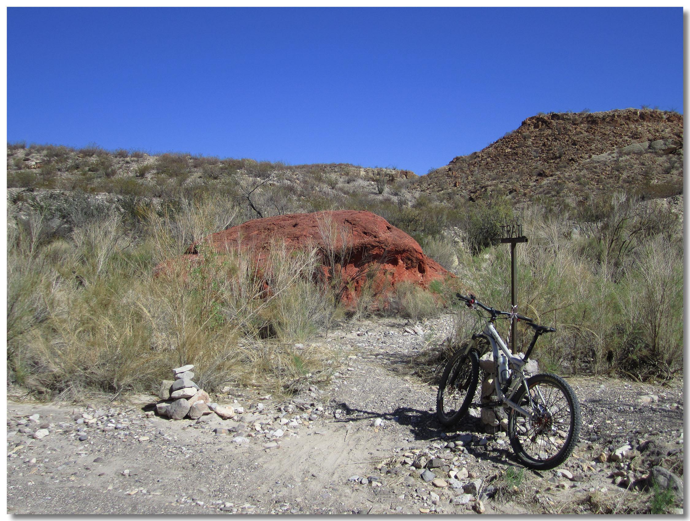 A mountain bike parked next to a trail sign amidst a rocky, arid landscape. In the background, a large red rock formation is visible, surrounded by sparse vegetation and blue skies. Small stacked stones are positioned at the base of the sign, indicating the trail's location. Big Bend Ranch State Park mountain bike trail.
