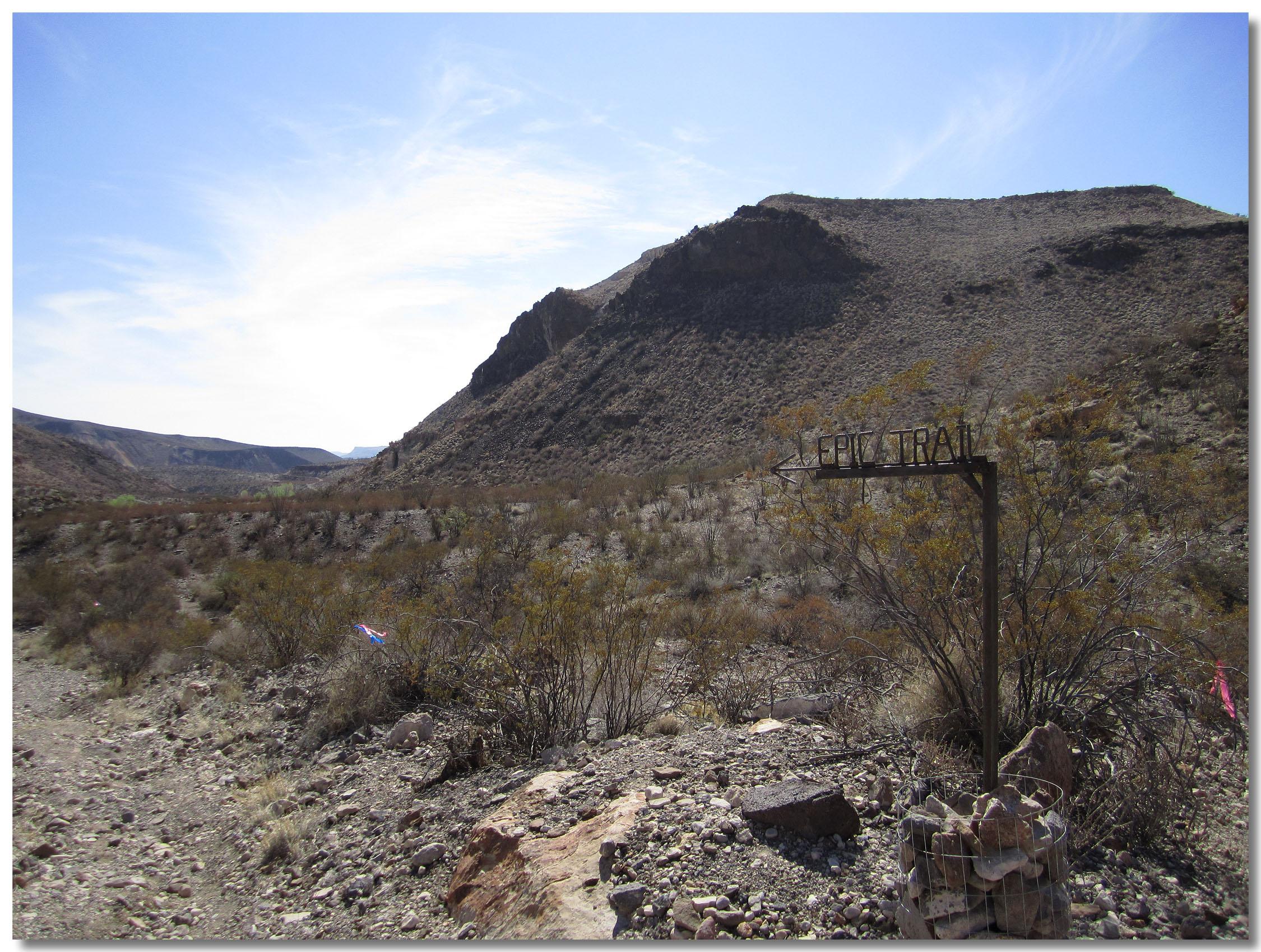 A desolate landscape featuring a rocky terrain, bushes, and rolling hills under a clear blue sky. A wooden sign marked "EPIZ TRAIL" points to the left, indicating the direction of the trail, while scattered rocks and a small stone pile are visible in the foreground. Big Bend Ranch State Park mountain bike trail.
