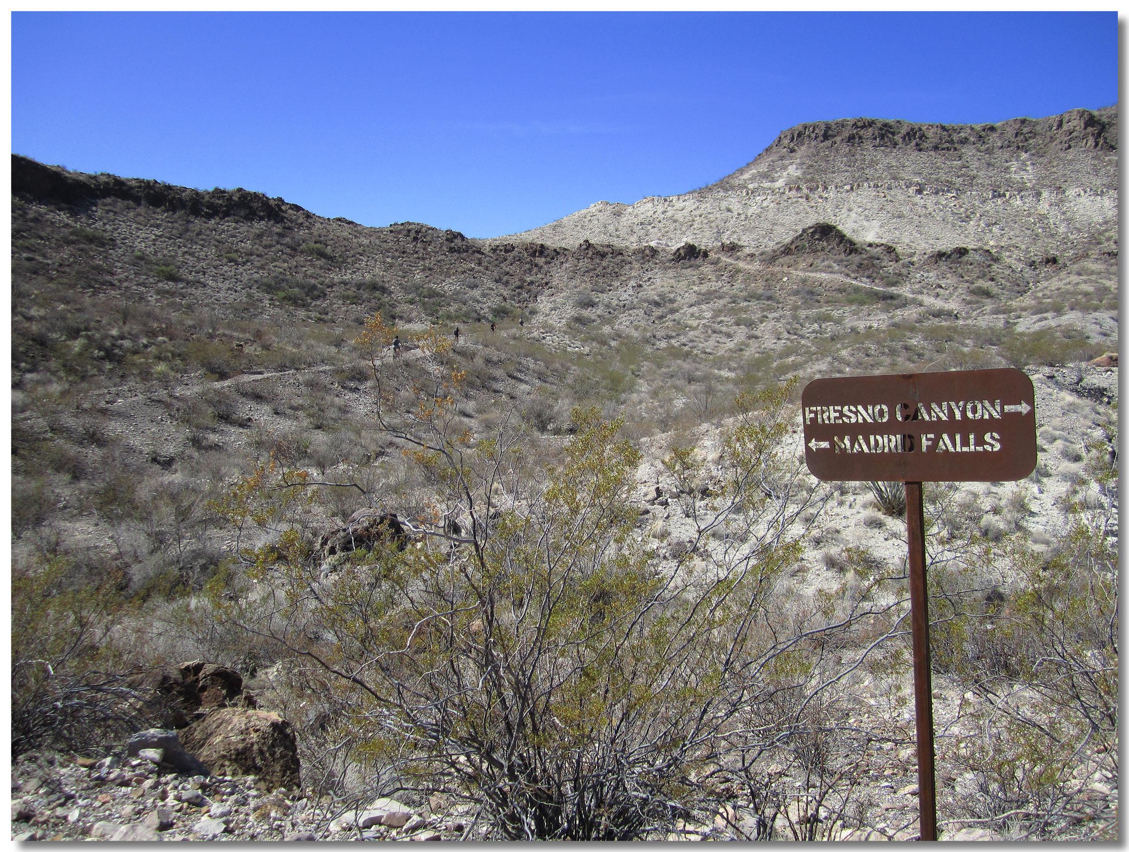 A signpost indicating directions to Fresno Canyon and Madrid Falls, set against a rocky, arid landscape with sparse vegetation and a clear blue sky. Big Bend Ranch State Park mountain bike trail.