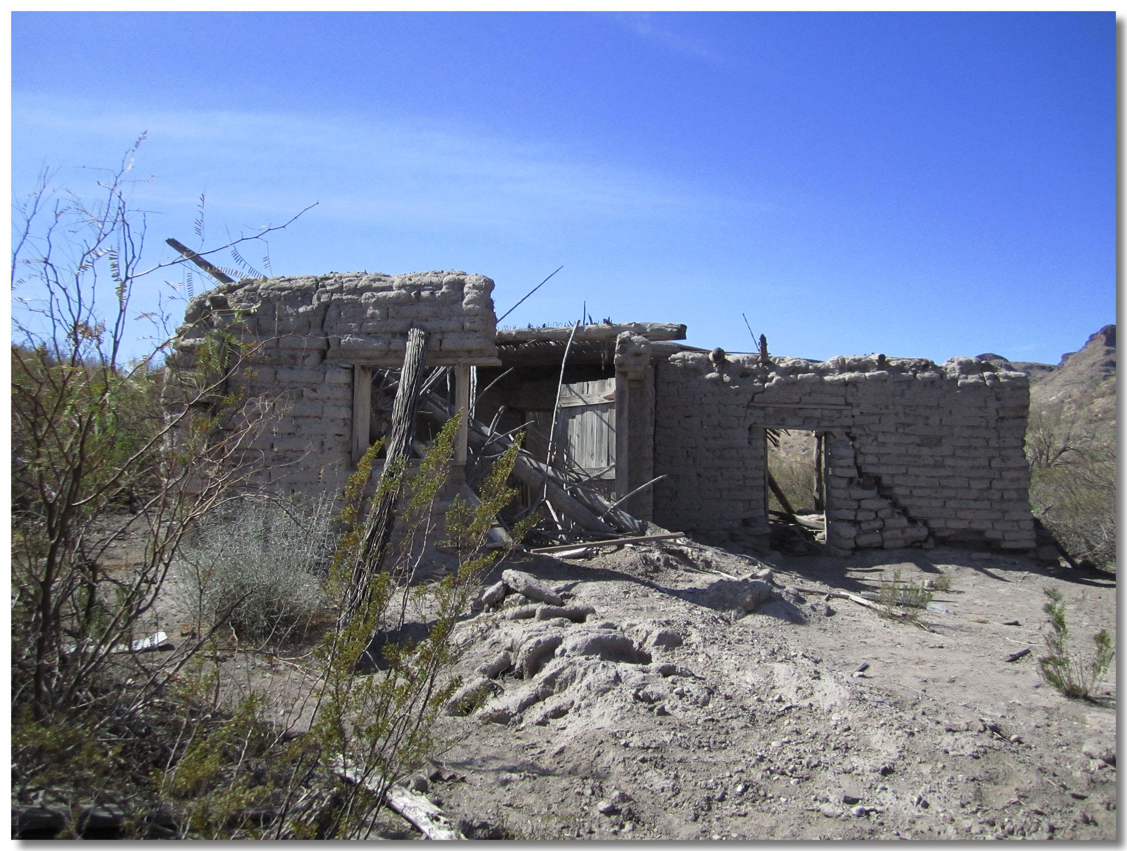 A weathered, abandoned building made of mud bricks sits in a dry landscape. The structure has crumbling walls and a partially collapsed roof, surrounded by sparse vegetation and gravelly ground under a clear blue sky. Big Bend Ranch State Park mountain bike trail.