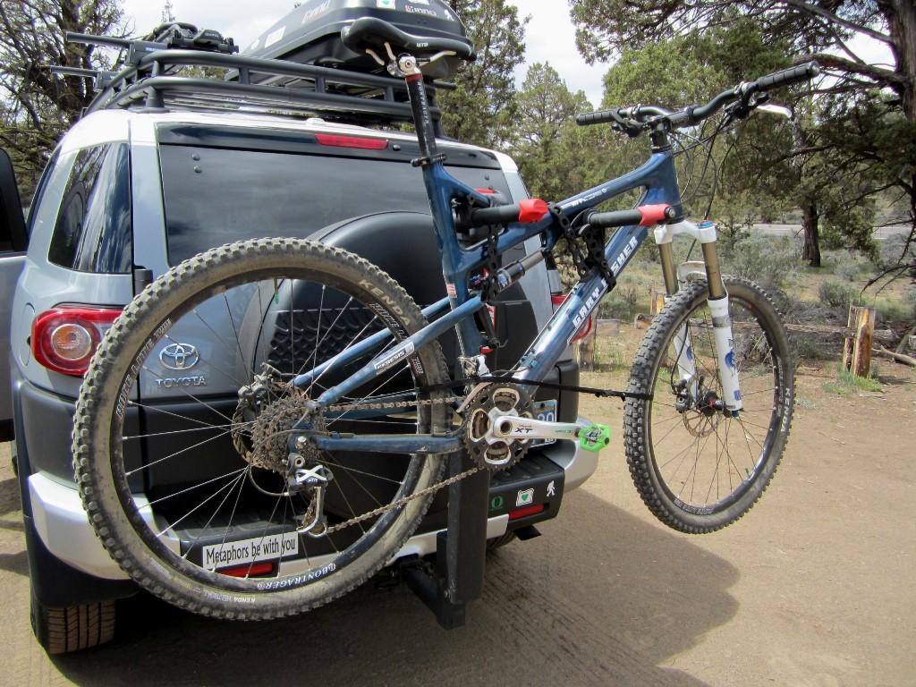 Gary Fisher HiFi Carbon: A blue mountain bike secured to the back of a silver Toyota SUV, parked in a natural outdoor setting. The bike is mounted on a rear bike rack, showing off its muddy tires and high-performance components. Trees and gravel can be seen in the background, suggesting a recreational area.