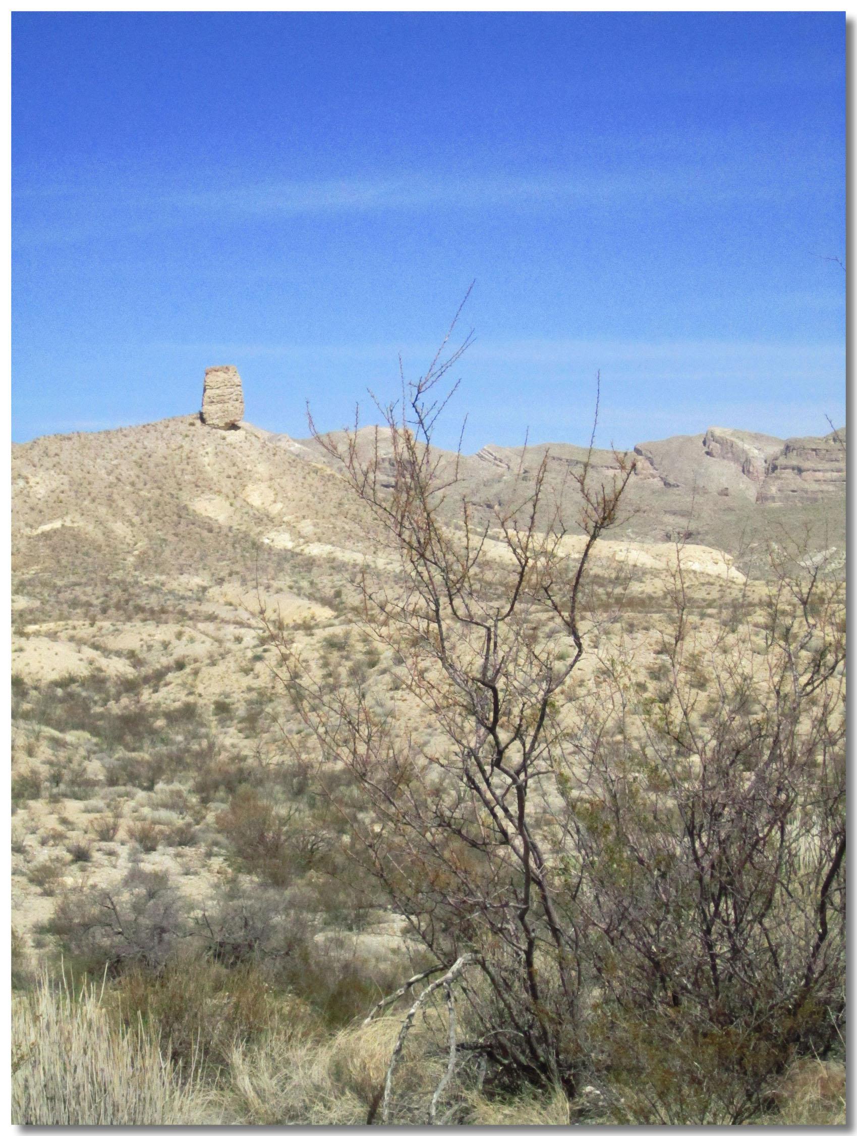 A rocky outcrop stands prominently on a hill, surrounded by a desert landscape featuring sparse vegetation and rolling hills beneath a clear blue sky. Big Bend Ranch State Park mountain bike trail.
