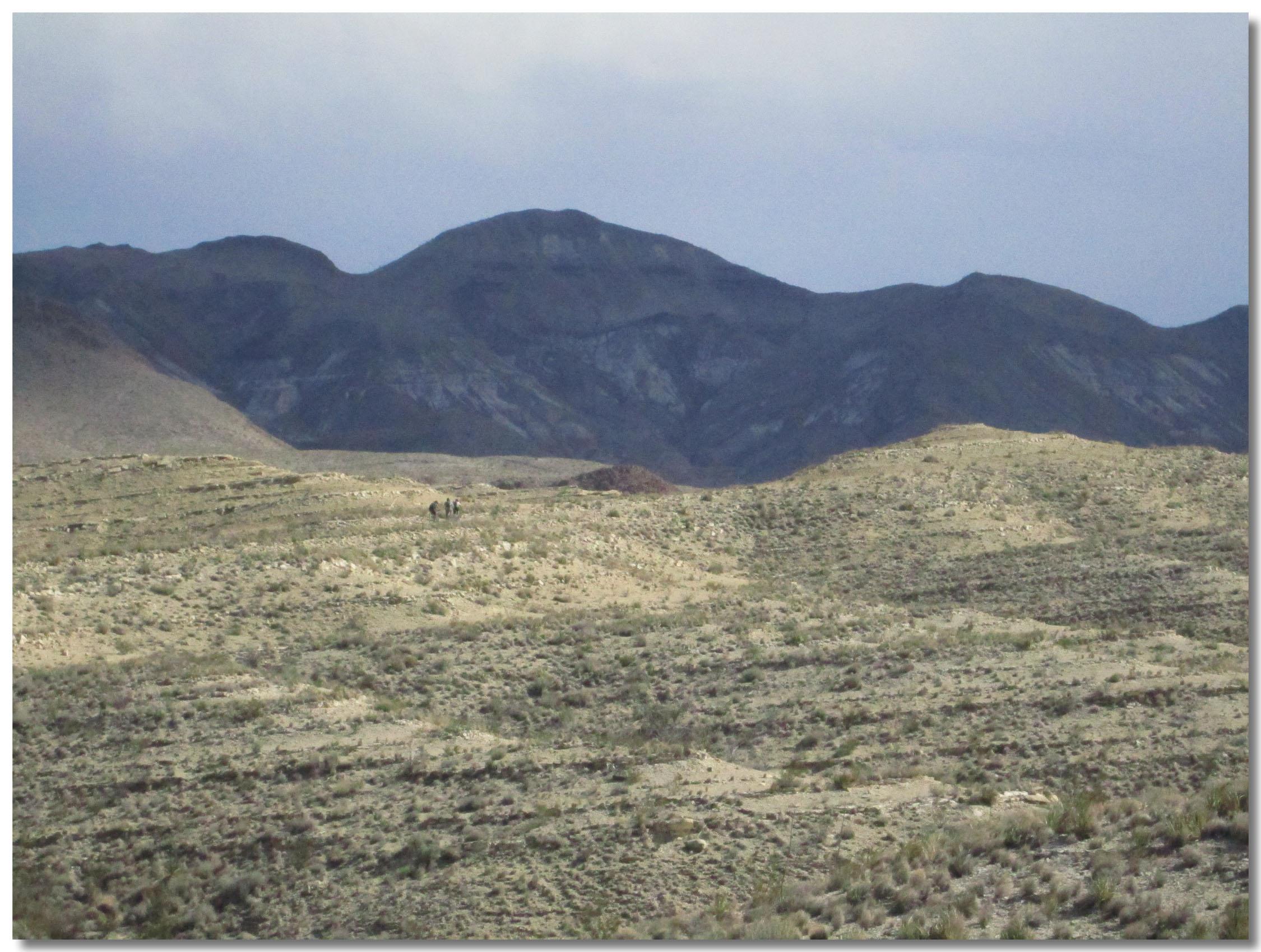 A panoramic view of a barren landscape featuring rolling hills and distant mountains under a cloudy sky. Sparse vegetation covers the ground, with a few figures visible in the distance, providing a sense of scale to the expansive scene. Big Bend State Park mountain bike trail.