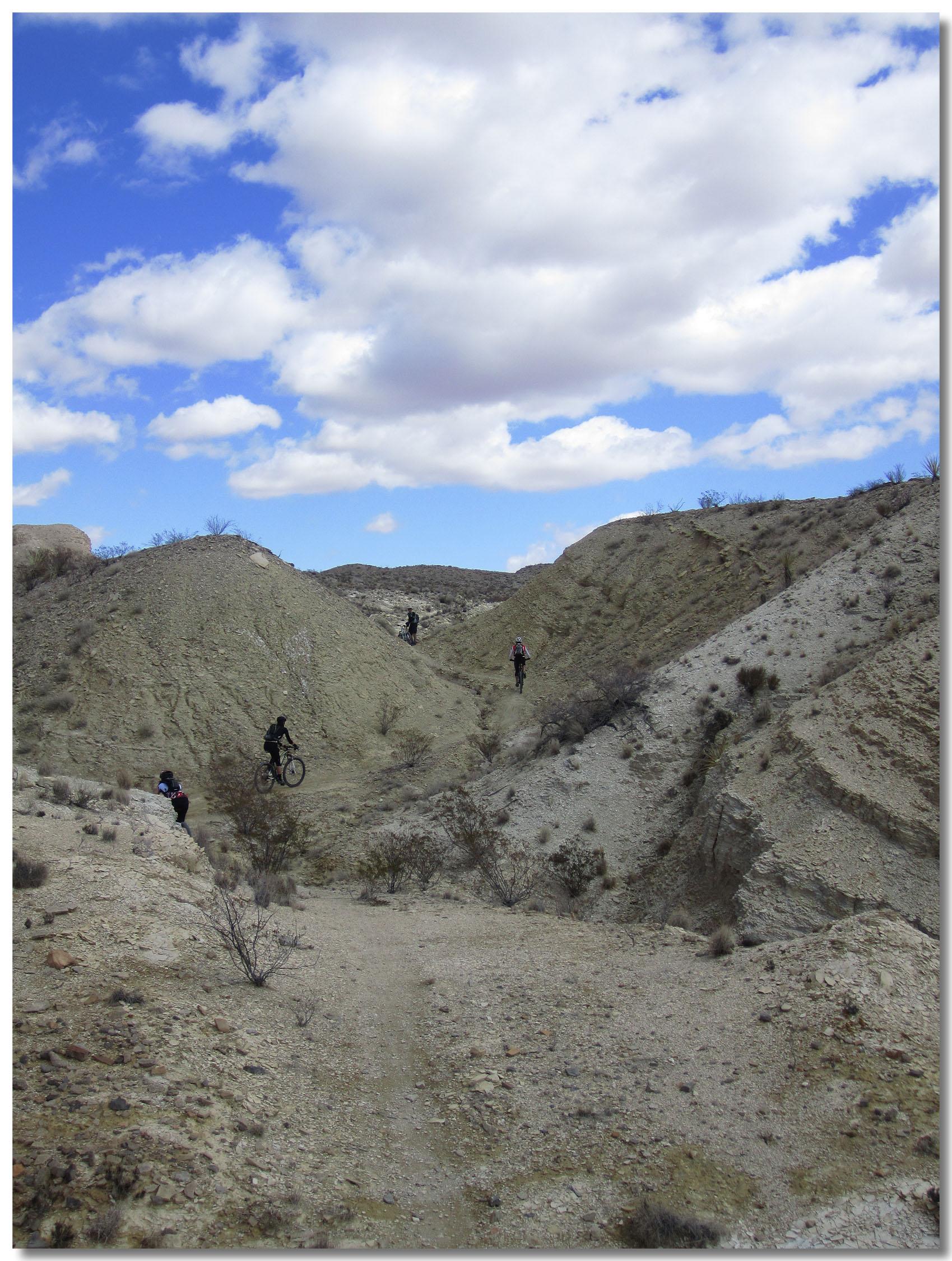 Bikers navigating a rugged terrain with sandy hills and sparse vegetation under a partly cloudy blue sky. Big Bend Ranch State Park mountain bike trail.
