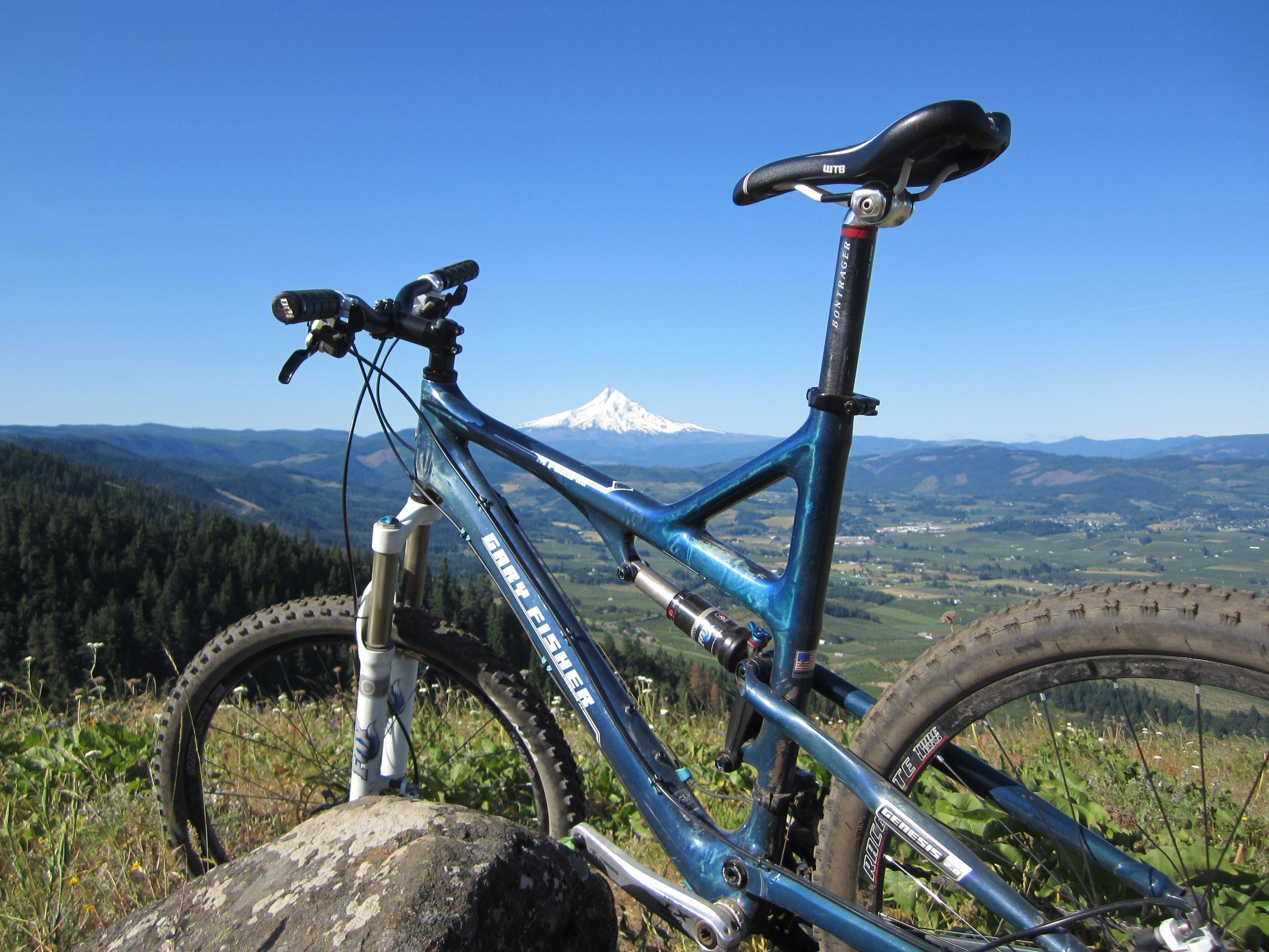A mountain bike resting on a rocky outcrop, with a panoramic view of rolling hills and a snow-capped mountain in the background under a clear blue sky. Whoopdee mountain bike trail.