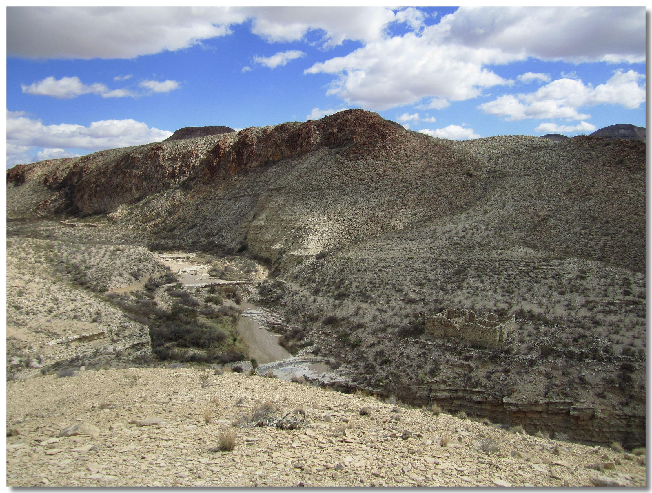 A panoramic view of a rugged desert landscape featuring arid hills, a winding river, and remnants of an old stone structure. The sky is partly cloudy, with a mix of blue and white clouds casting shadows on the terrain. Sparse vegetation, including shrubs and grasses, dot the rocky ground. Big Bend Ranch State Park mountain bike trail.