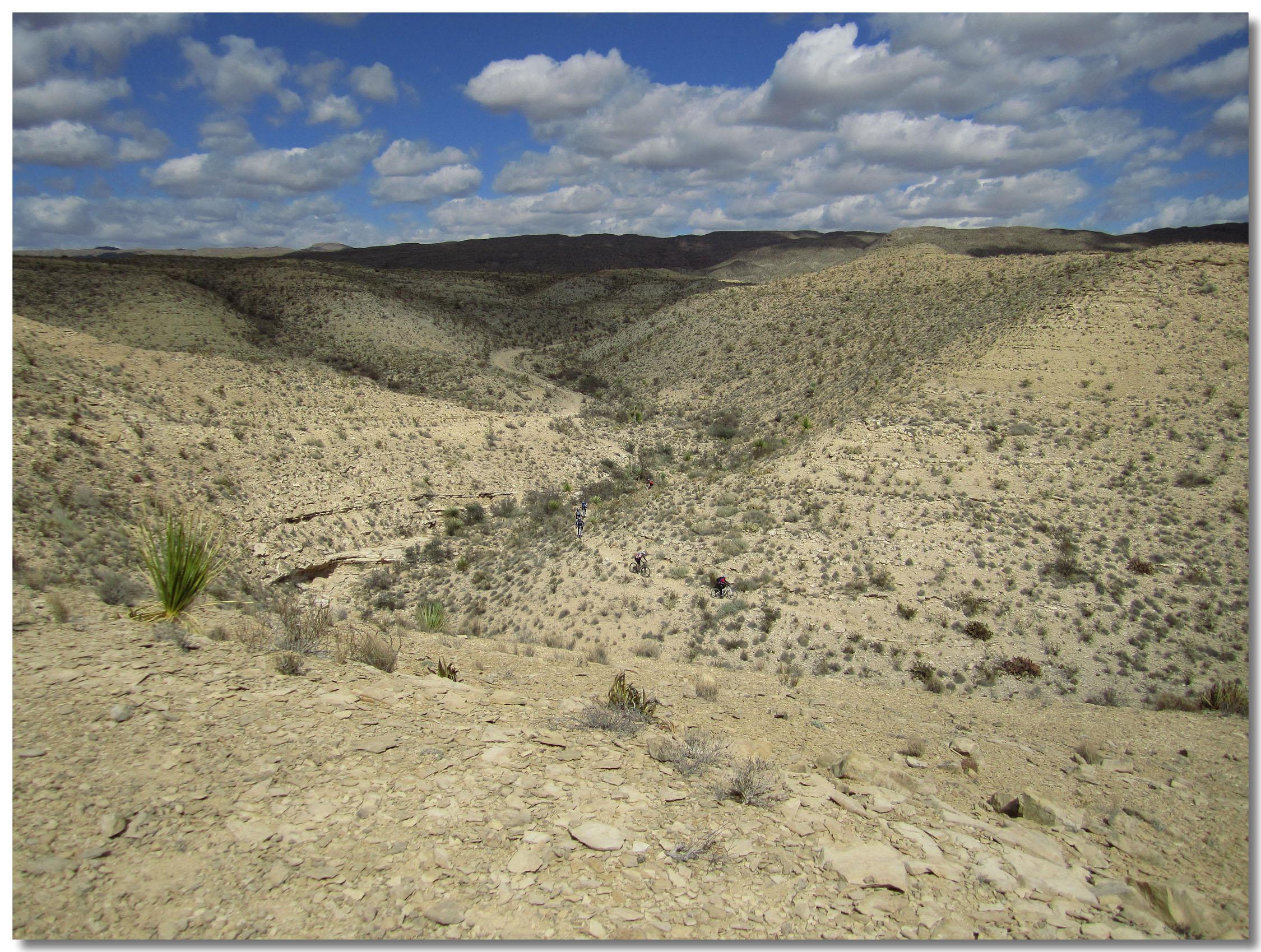 A wide view of a dry, desert landscape featuring rolling hills and a winding pathway. The terrain is mostly bare with sparse vegetation, including low shrubs and a few prominent plants. The sky is partly cloudy, showcasing a mix of blue and white clouds. The image captures the natural beauty of a remote, arid environment. Big Bend State Park mountain bike trail.