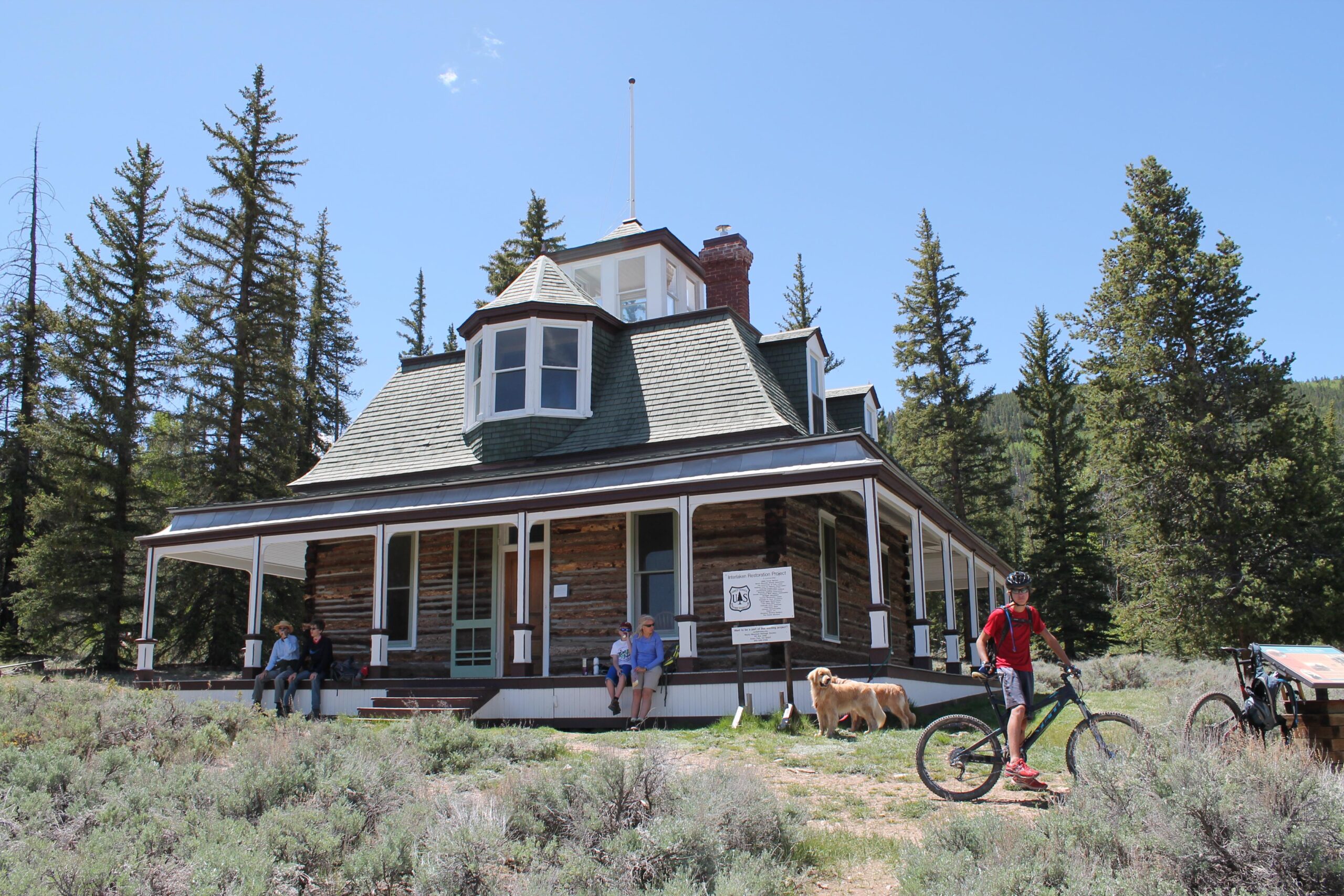 A historic log cabin in a forested area, featuring a wraparound porch and a distinctive turret. Two people sit on the porch, while another person in a red shirt stands next to a mountain bike. A golden retriever lies on the grass nearby, and tall trees surround the cabin under a clear blue sky. Twin Lakes Loop mountain bike trail.
