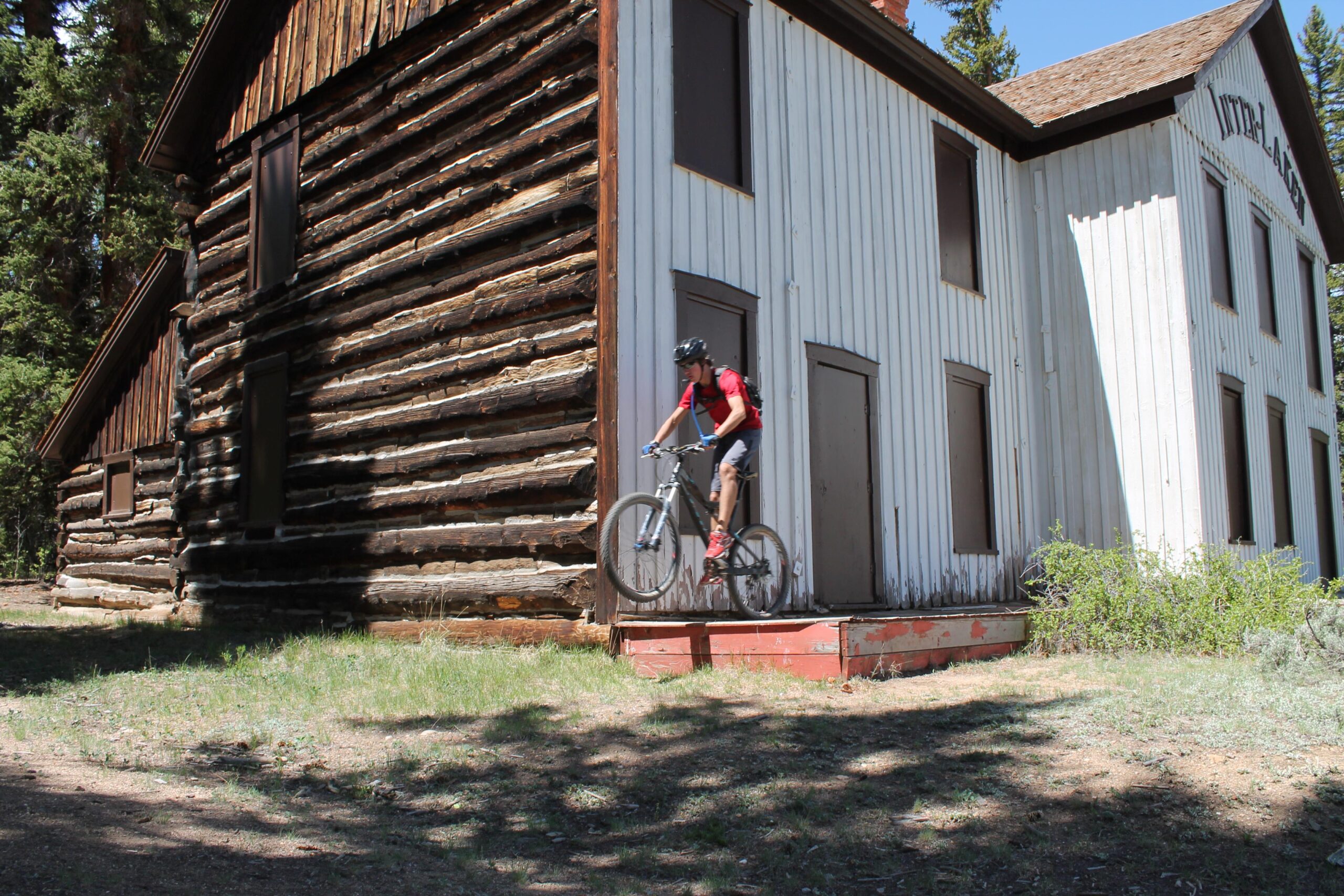 A person wearing a helmet and riding a mountain bike is jumping off a small wooden platform in front of a rustic, two-tone log cabin with a white and dark brown exterior. The cabin is surrounded by greenery and trees, and the scene is set on a sunny day. Twin Lakes Loop mountain bike trail.