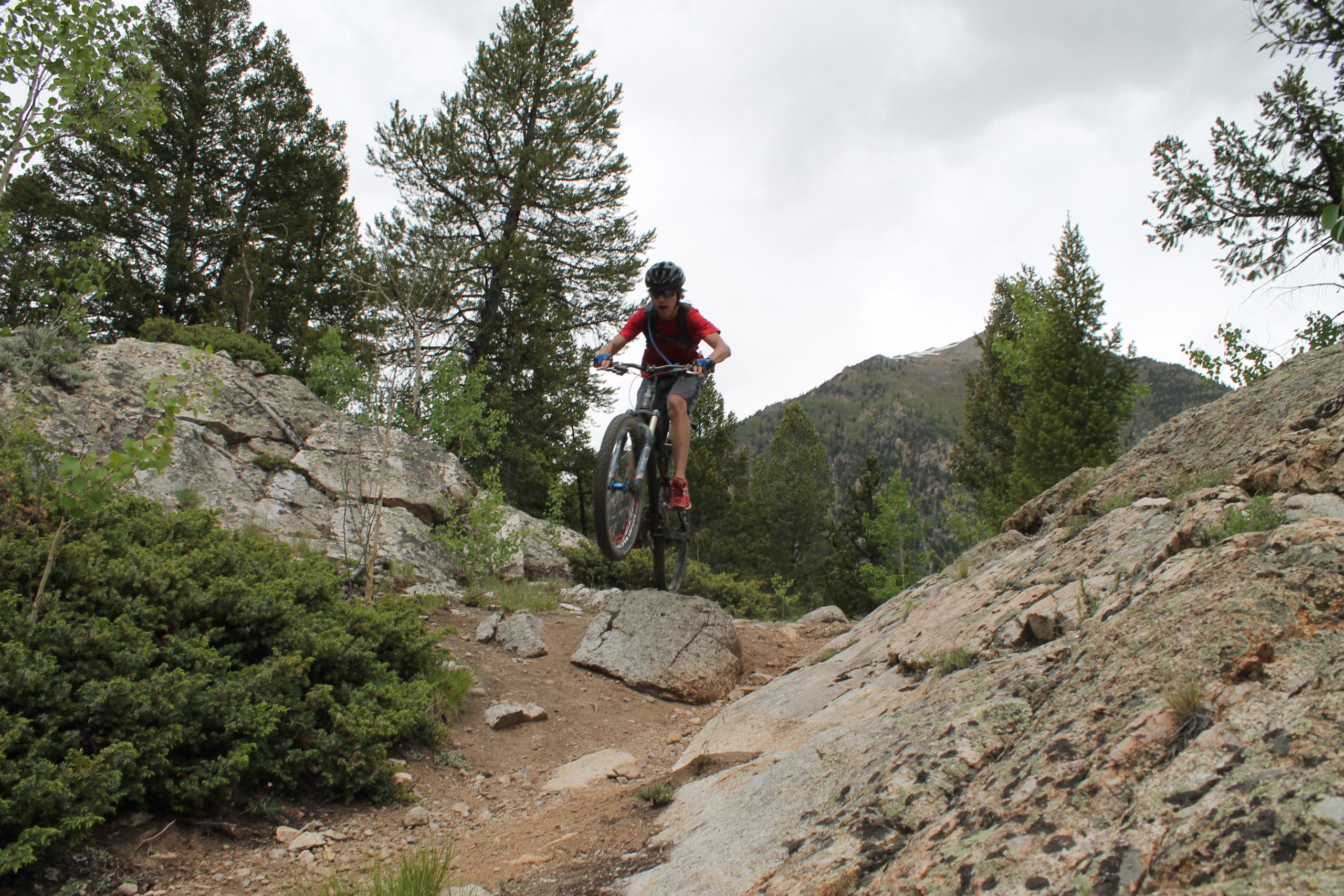 A mountain biker in a red shirt and helmet performs a jump over a rock on a rugged trail surrounded by trees and mountains under a cloudy sky. Twin Lakes Loop mountain bike trail.