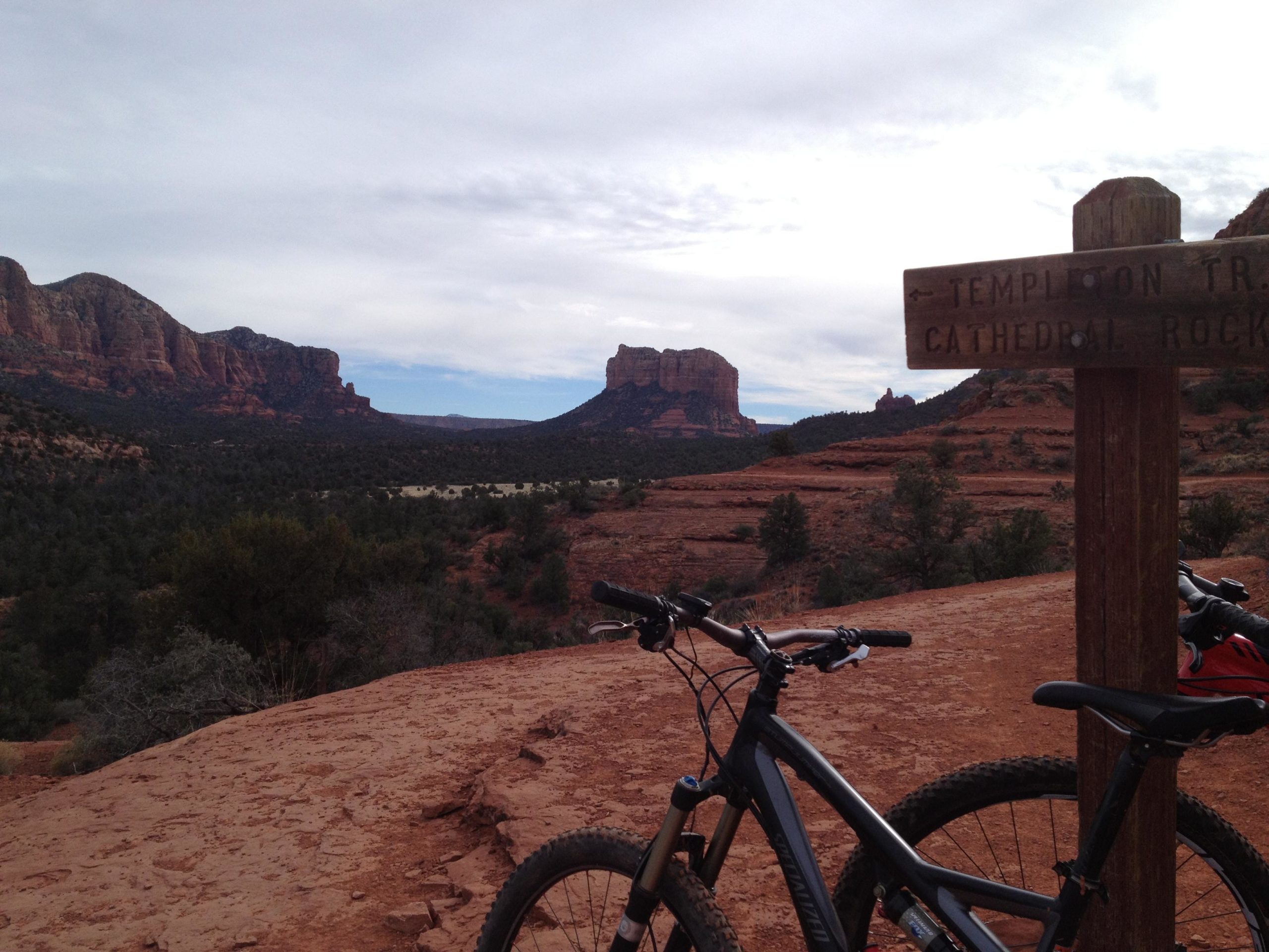 A scenic view of red rock formations in a desert landscape, featuring a wooden signpost indicating the direction to "Templton Trail" and "Cathedral Rocks." In the foreground, two mountain bikes are parked on the rocky terrain, with vegetation partially visible, under a cloudy sky. Templeton mountain bike trail.